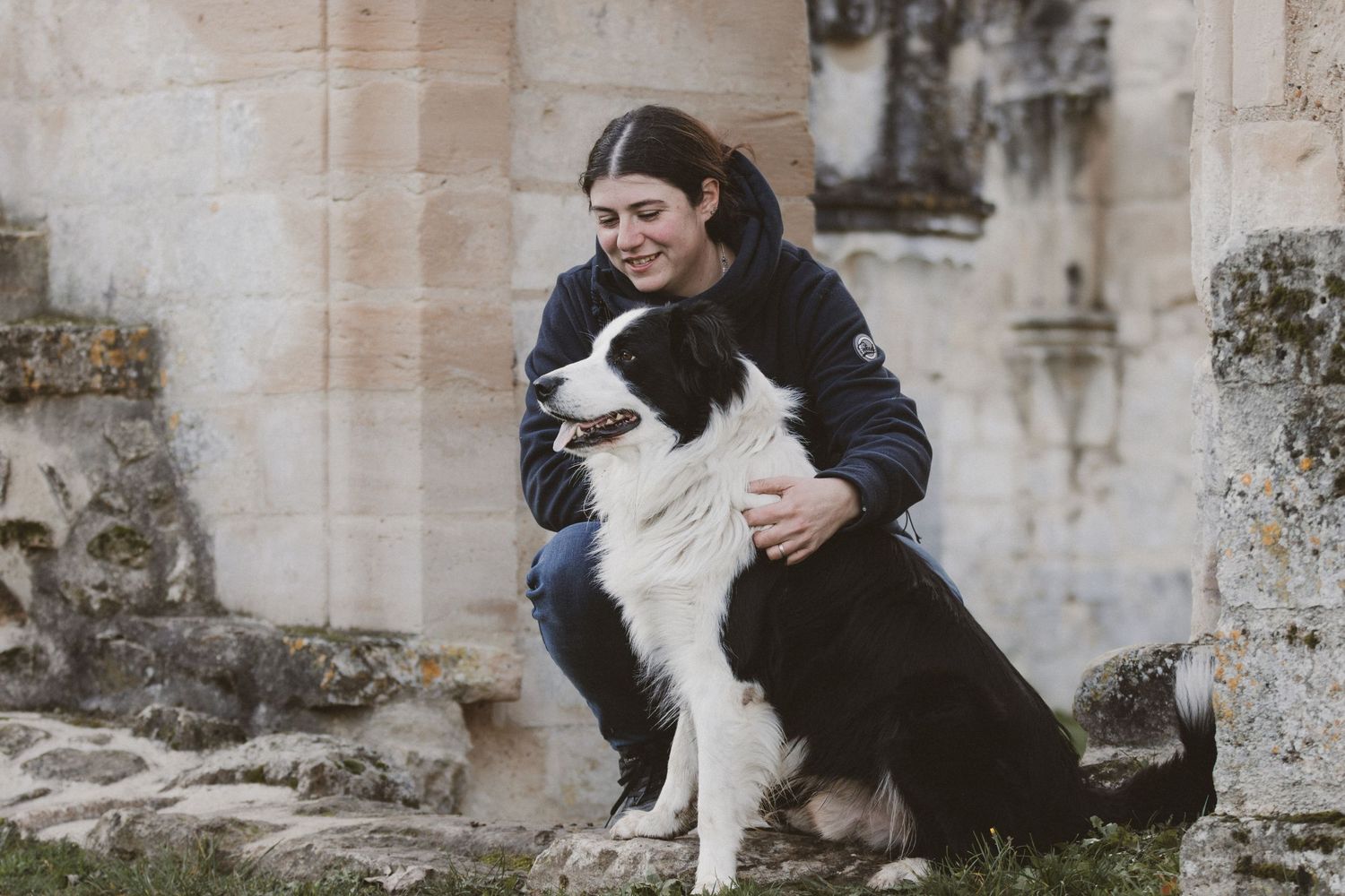 Smiling woman crouching beside a black and white border collie dog near a stone building.
