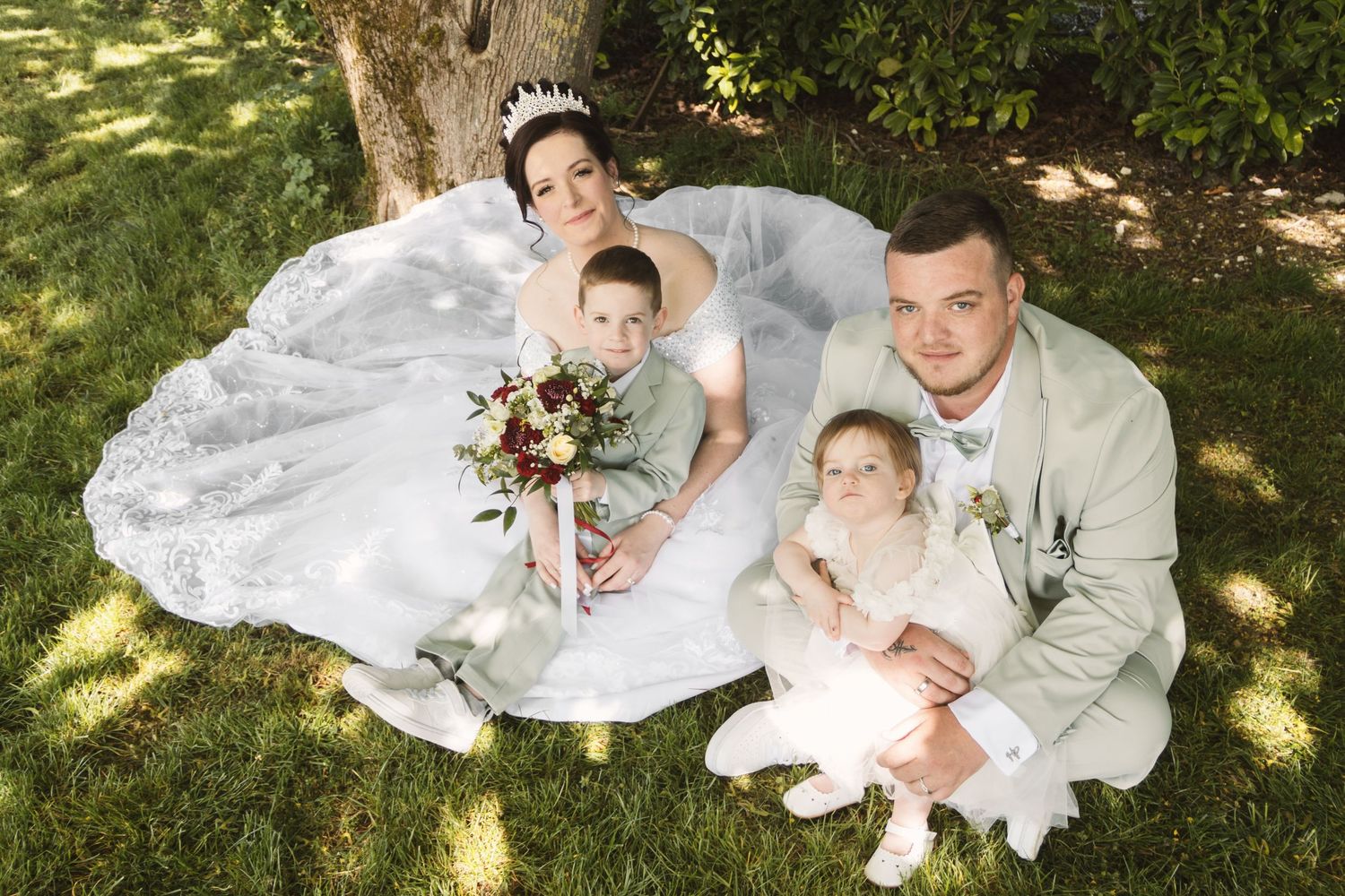 Bride and groom sit on grass with two young children, bride in white gown holding bouquet, groom in sage suit.