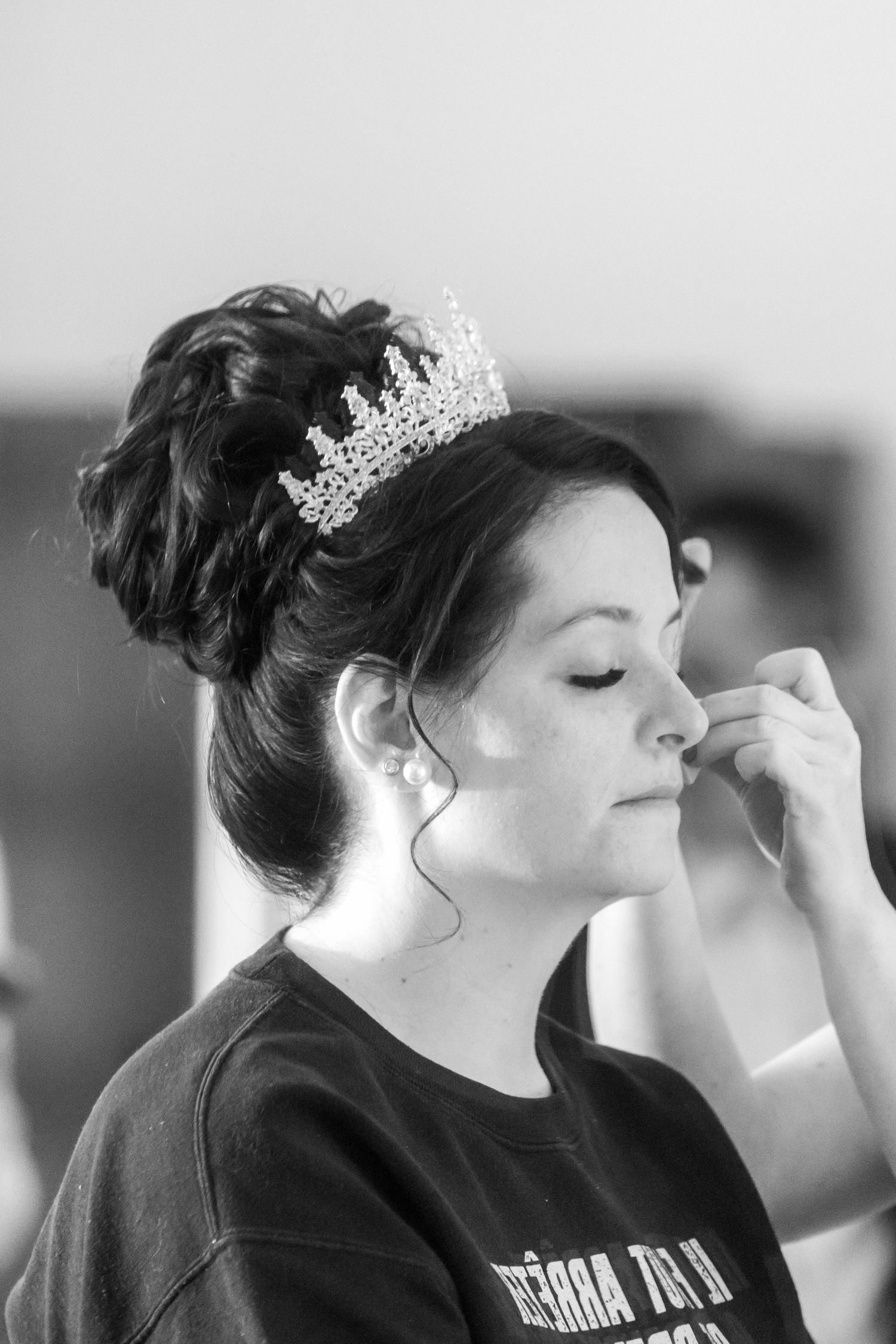 Black and white photo of a bride wearing a tiara with an updo hairstyle getting makeup applied before her wedding.