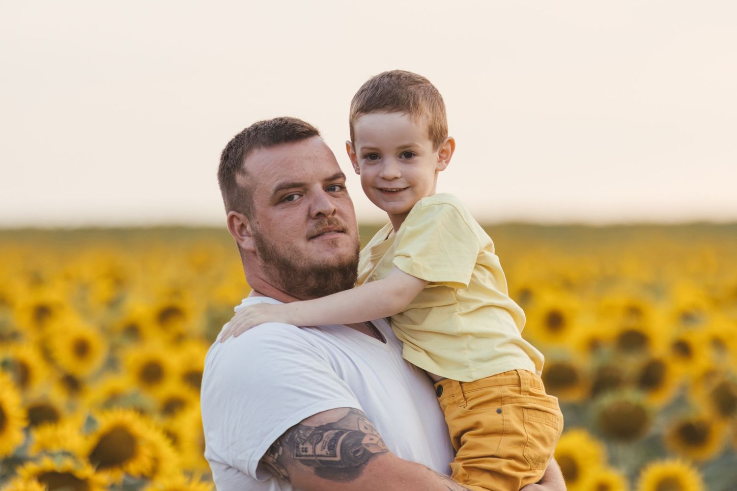 A tattooed man holds a young boy in a sunflower field at sunset, both smiling warmly at the camera.