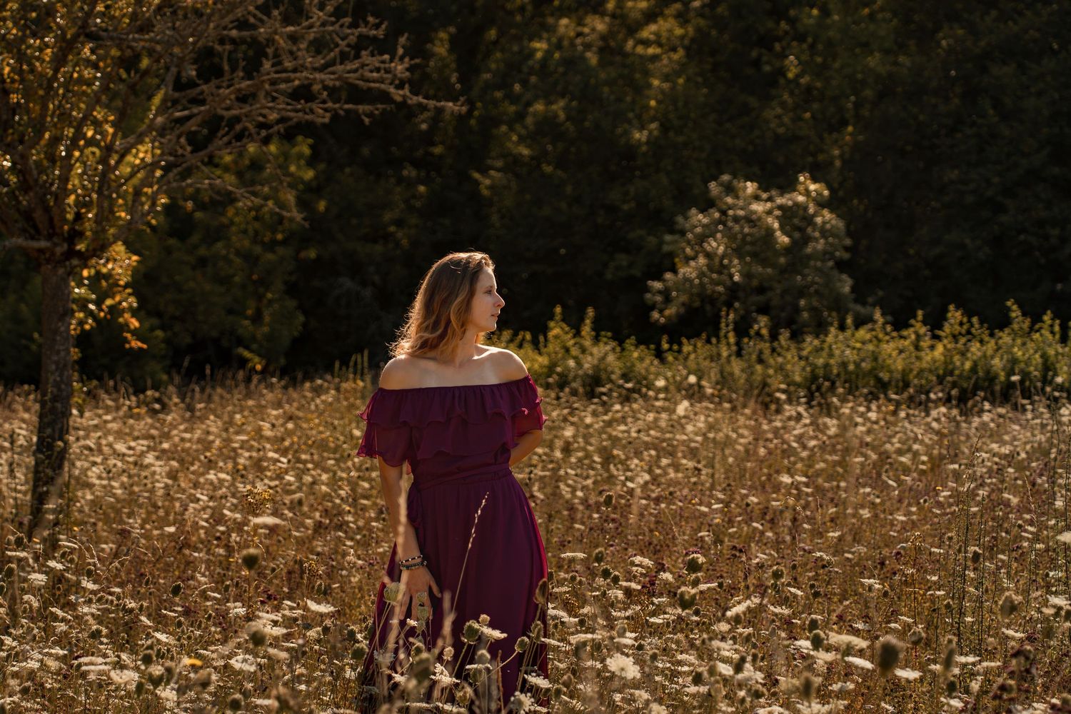 Woman in burgundy off-shoulder dress standing in golden wildflower field with autumn trees in background.