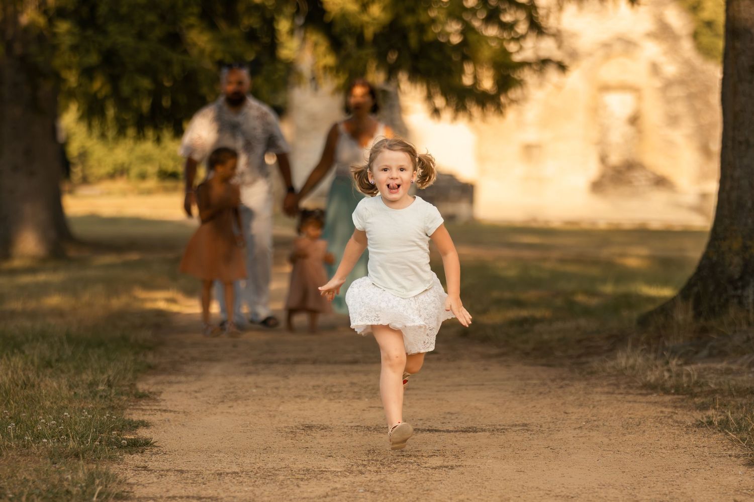 Young girl in white dress running joyfully toward camera on dirt path with family walking behind in golden sunlight.