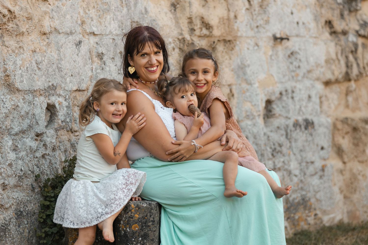 A mother with three young daughters poses together smiling against a stone wall outdoors.