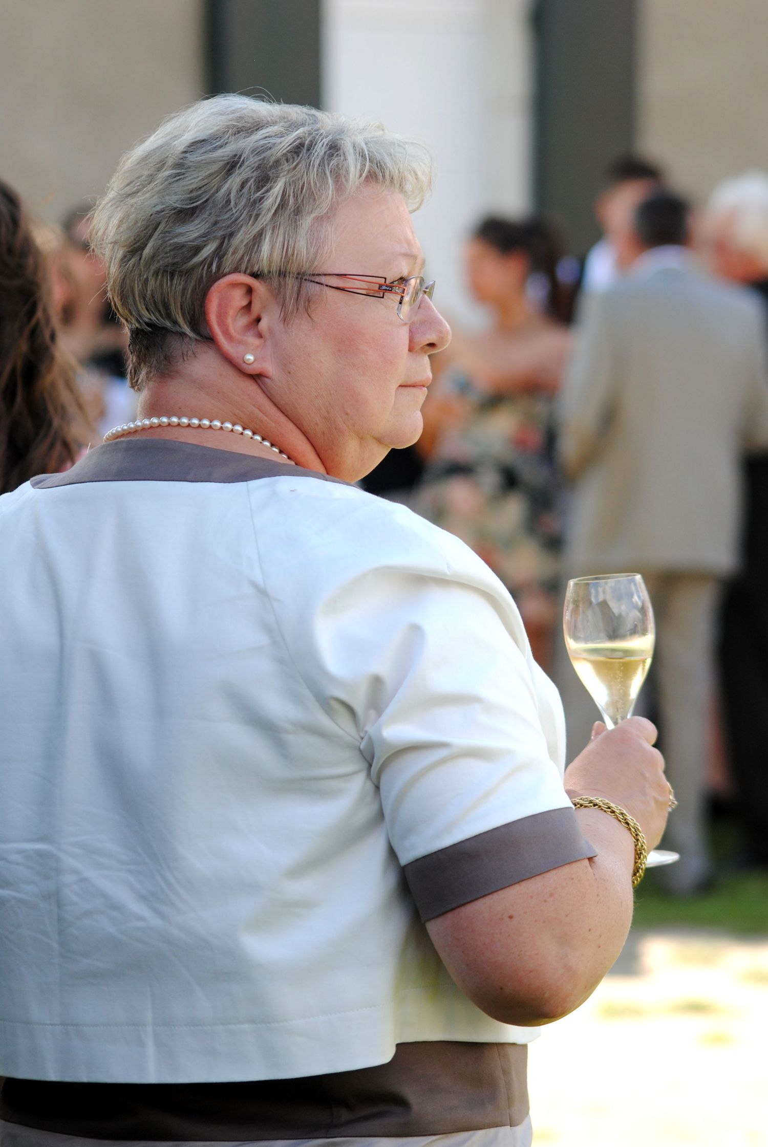 Older woman with gray hair holding a glass of white wine at an outdoor social gathering event.