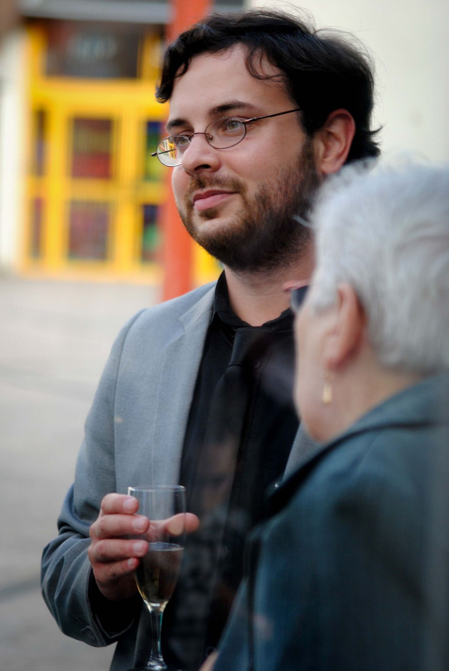 Bearded man with glasses holding a champagne glass in conversation at an outdoor social event.