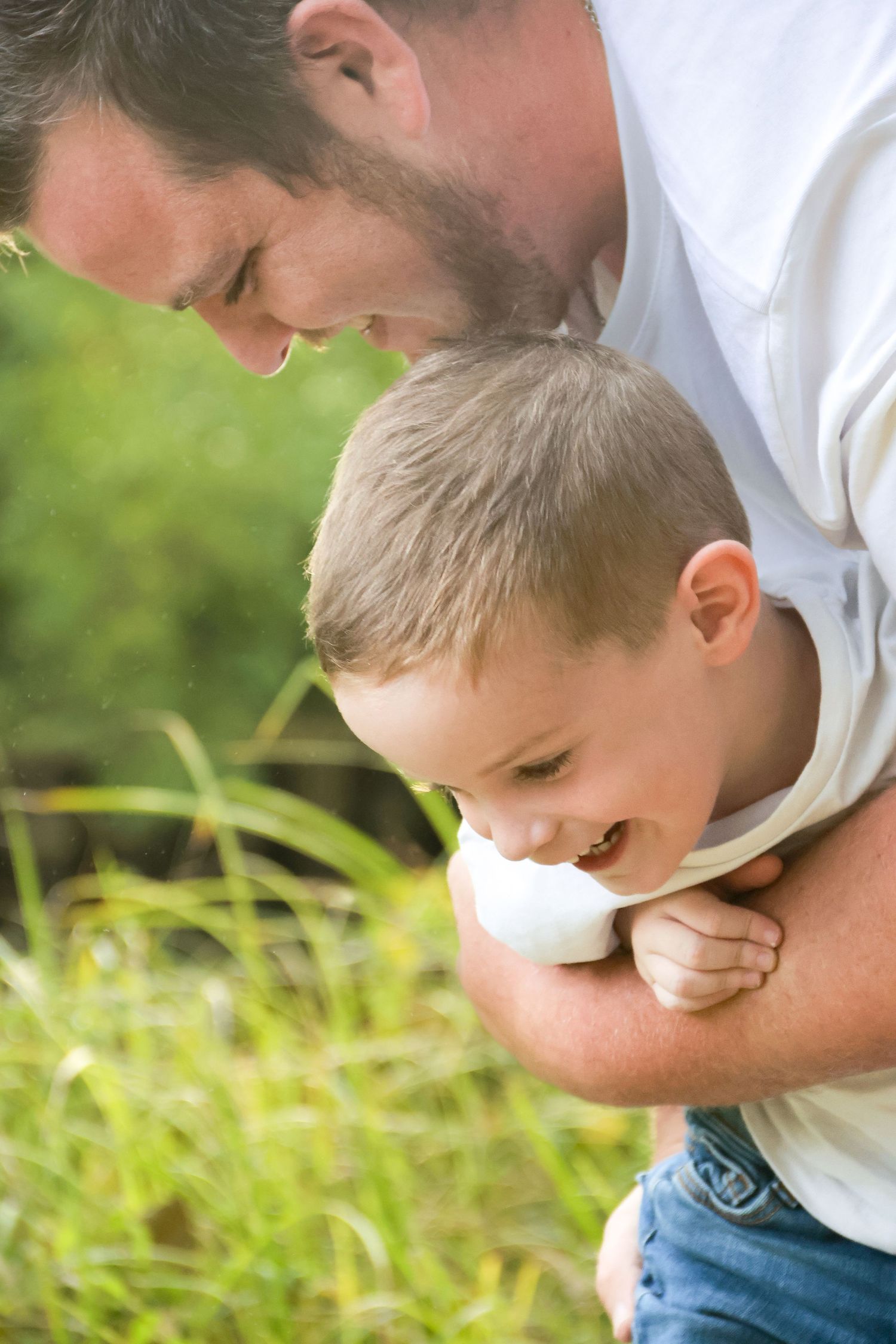 Father playfully hugging and tickling his laughing young son outdoors in a grassy green setting.