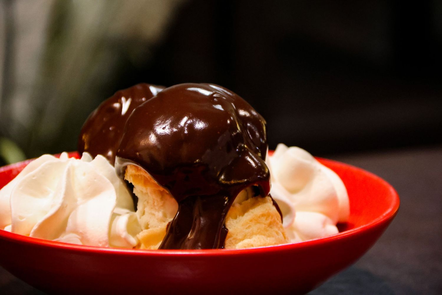 Red bowl filled with cream puffs topped with vanilla ice cream and rich chocolate fudge sauce.