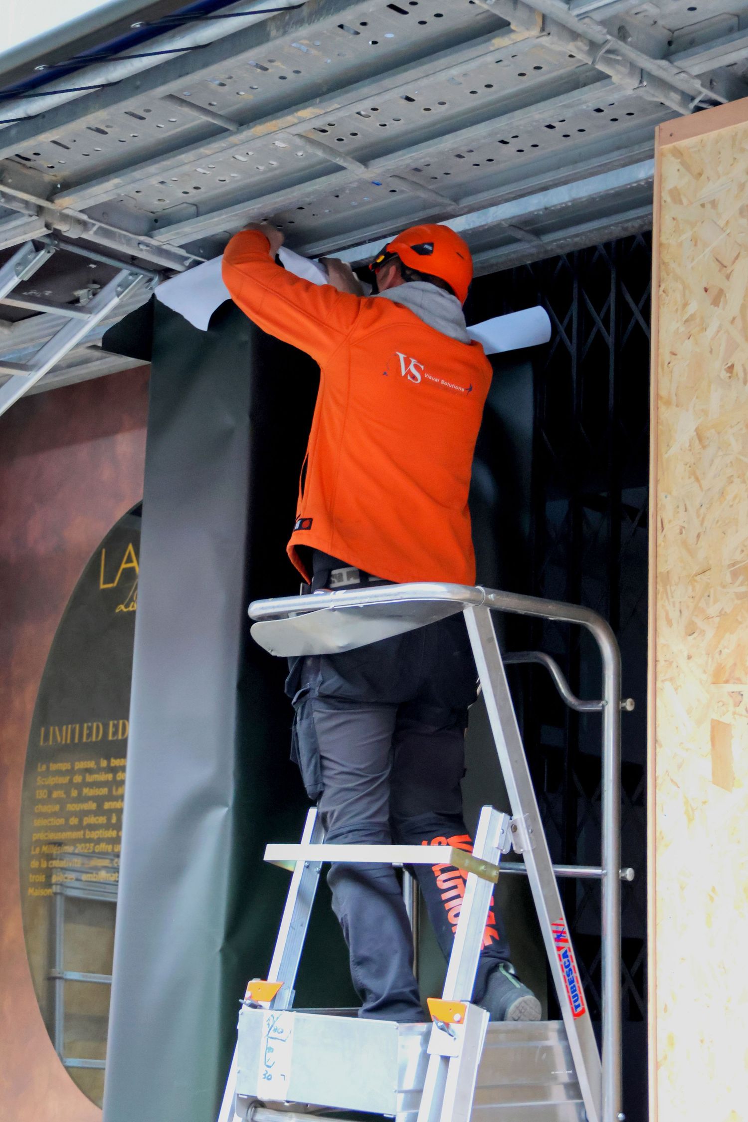 Worker in orange jacket on ladder installing ceiling panels at a construction or exhibition site.
