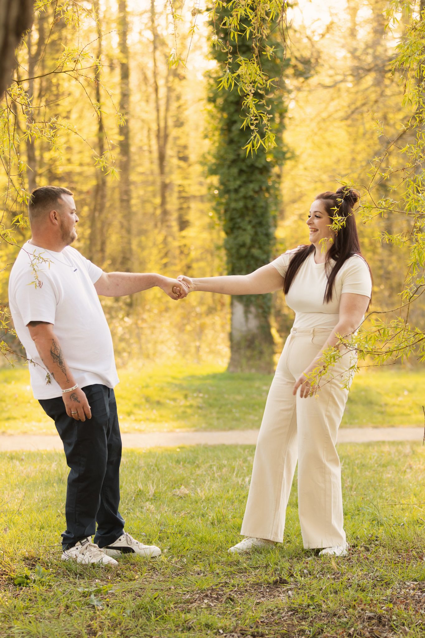 Couple holding hands and smiling in a sunny park with lush green trees and golden foliage in the background.