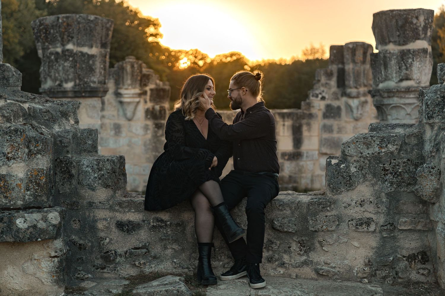 Couple in black outfits sitting intimately among ancient stone ruins at golden sunset.