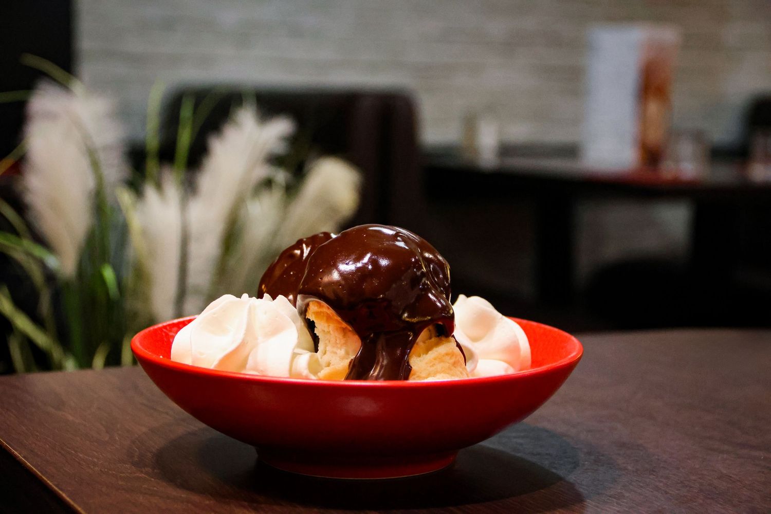 A red bowl with a banana split topped with chocolate sauce, ice cream, and whipped cream on a wooden table.