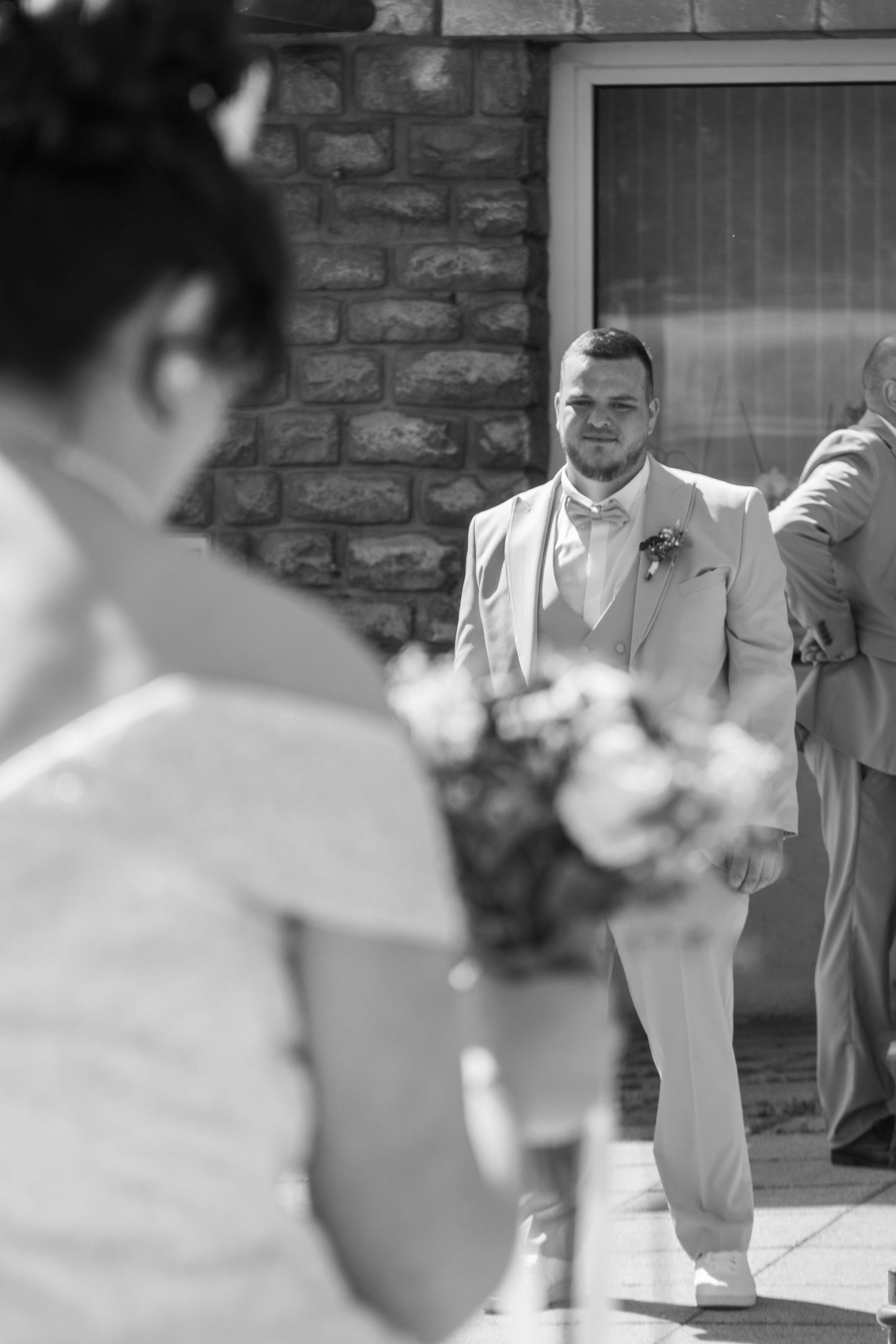 Black and white wedding photo of groom in light suit smiling at bride holding bouquet during outdoor ceremony.