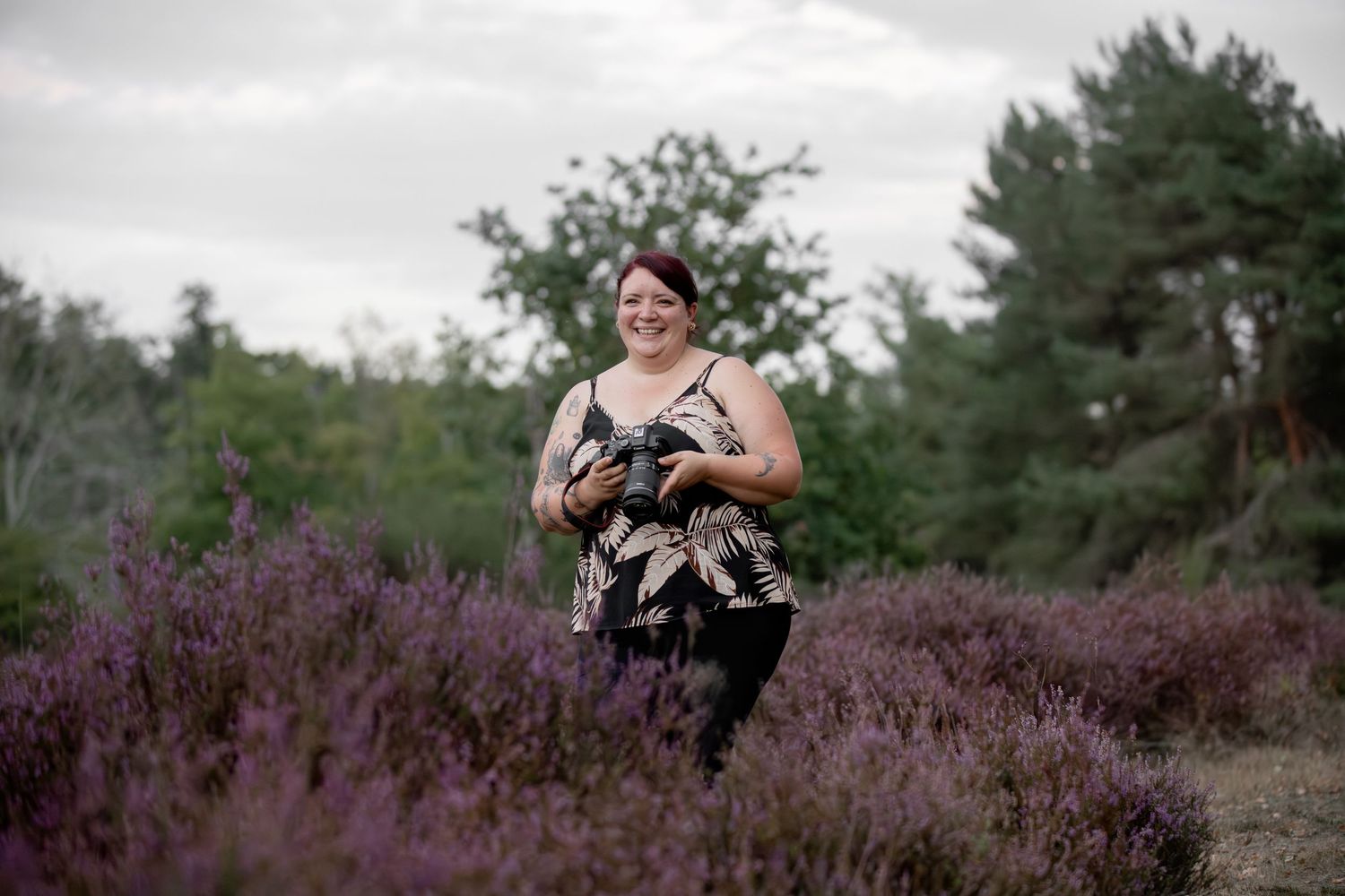 A cheerful red-haired woman holds a professional camera while walking through a purple heather field.