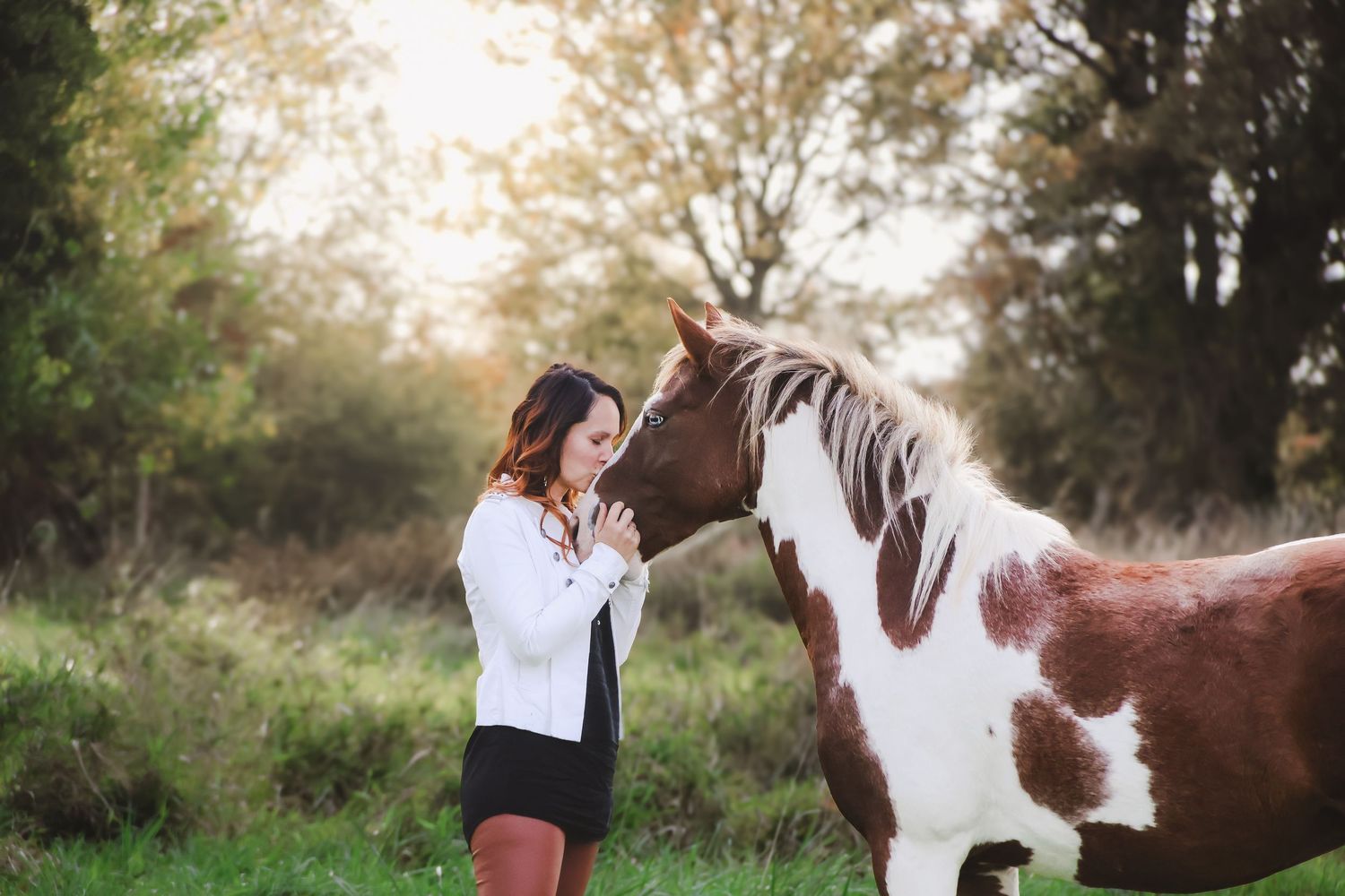 Young woman in white blouse tenderly bonding with a brown and white paint horse in a scenic outdoor setting.