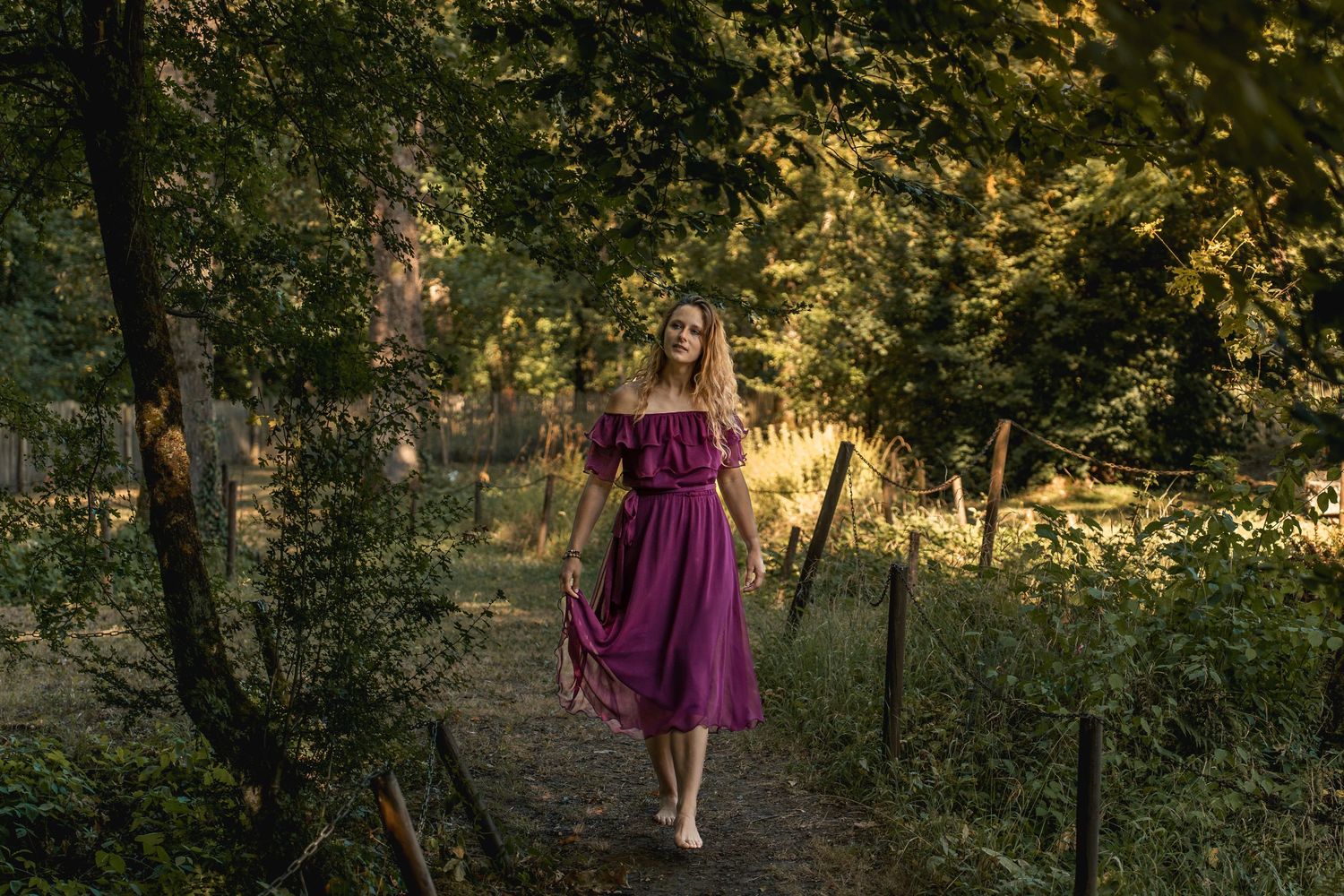 A woman in a purple off-shoulder midi dress walks along a sunlit forest path surrounded by lush green trees.