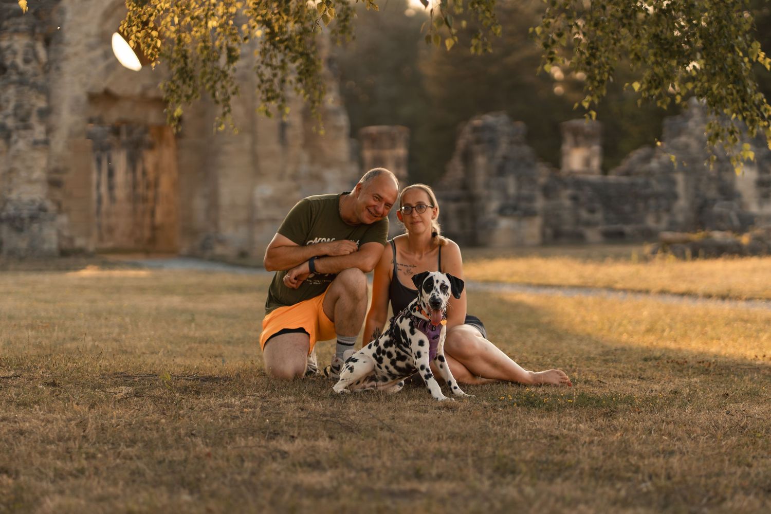 Couple sitting on grass with Dalmatian dog near ancient ruins during golden hour sunset.