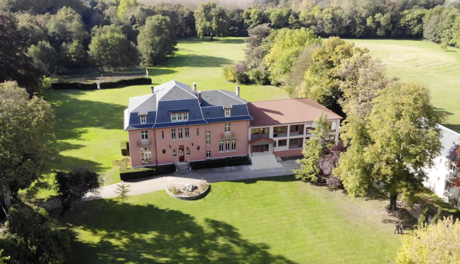 Aerial view of a grand pink manor house surrounded by lush green lawns and mature trees in a countryside setting.