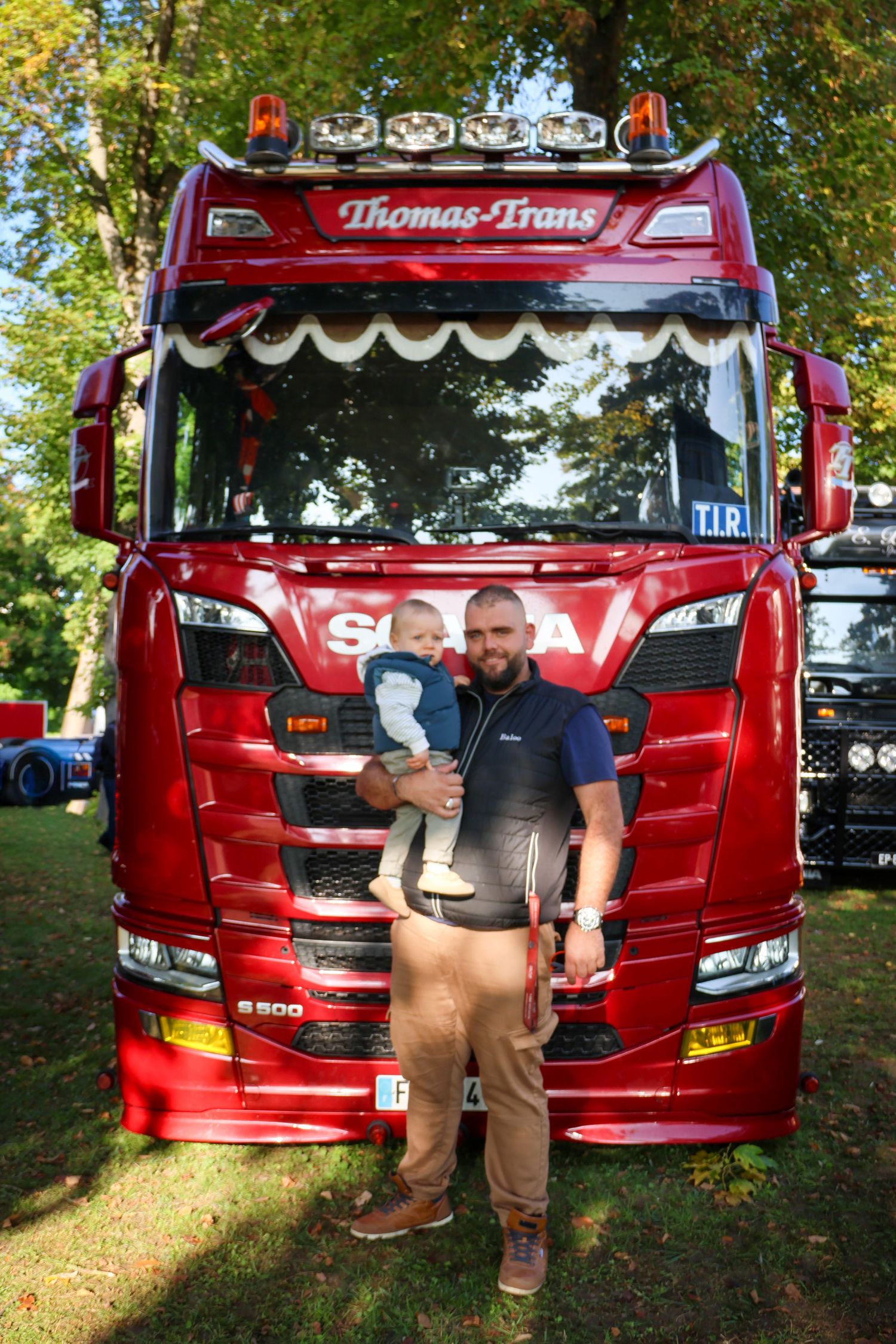 Man holding a baby poses proudly in front of a polished red Scania S500 Thomas-Trans truck.