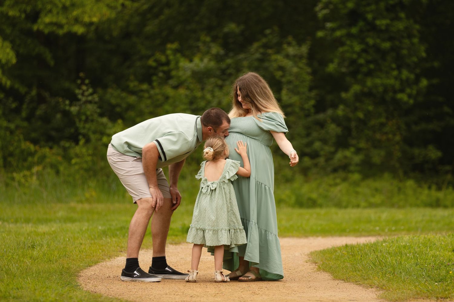 Pregnant woman in green dress with husband and toddler daughter on a winding park path surrounded by lush trees.