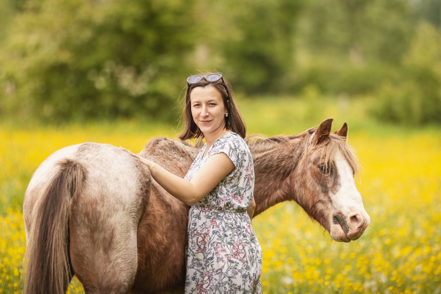 Woman in floral dress posing with a brown and white horse in a sunny yellow wildflower field.