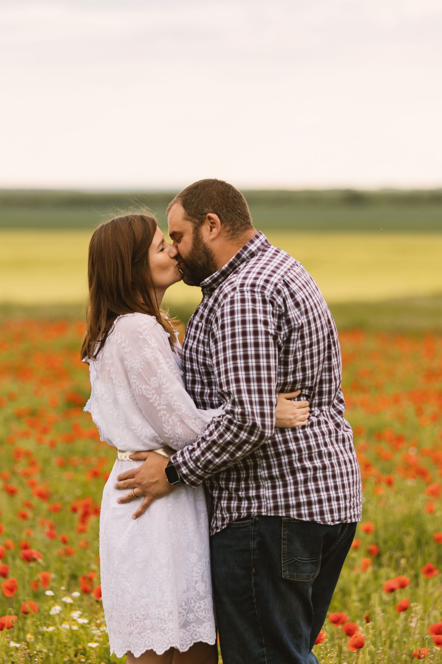 Couple sharing a romantic kiss in a vibrant red poppy field, woman in white dress, man in plaid shirt.