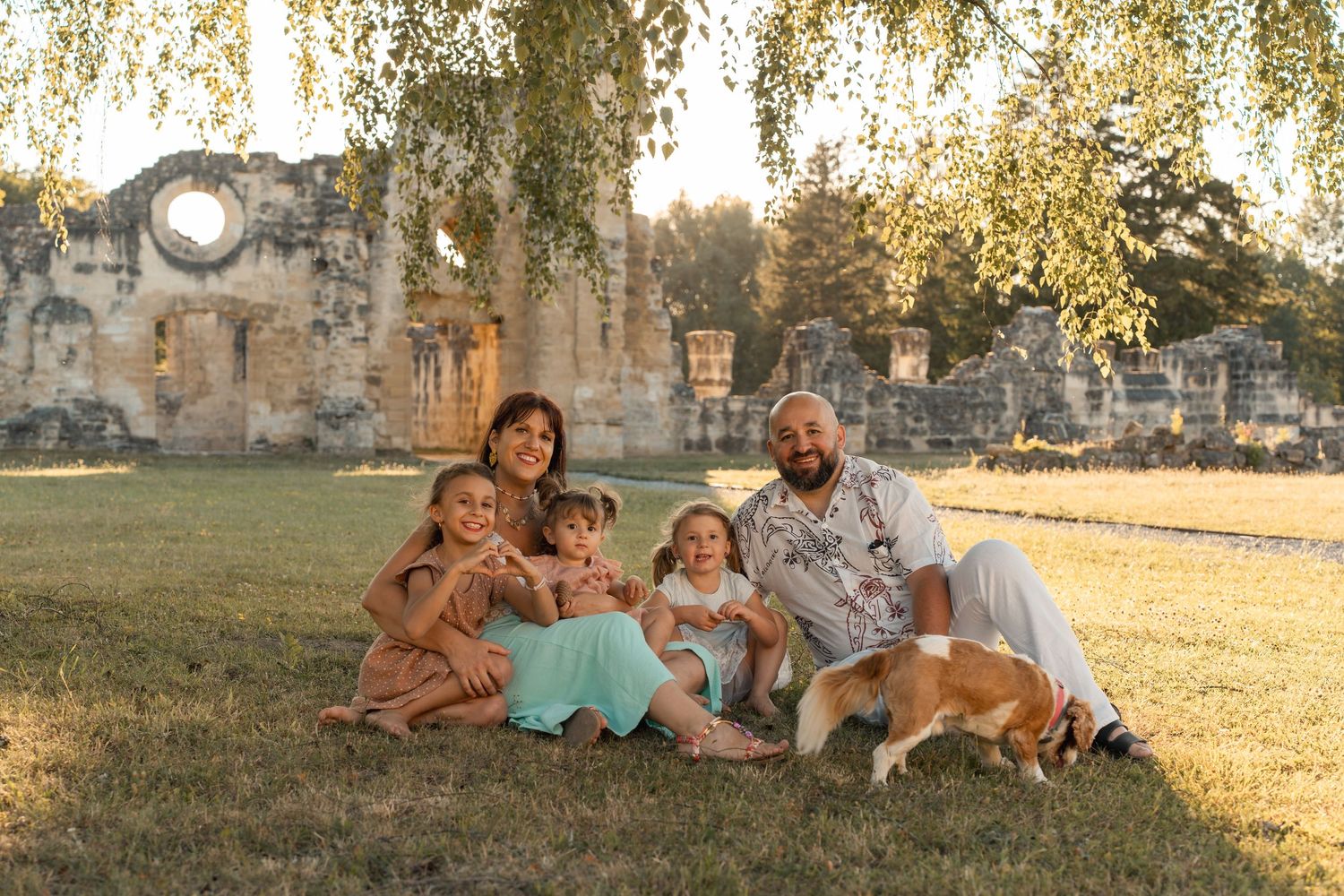 Family of four with dog sitting on grass near ancient stone ruins during golden hour sunset.