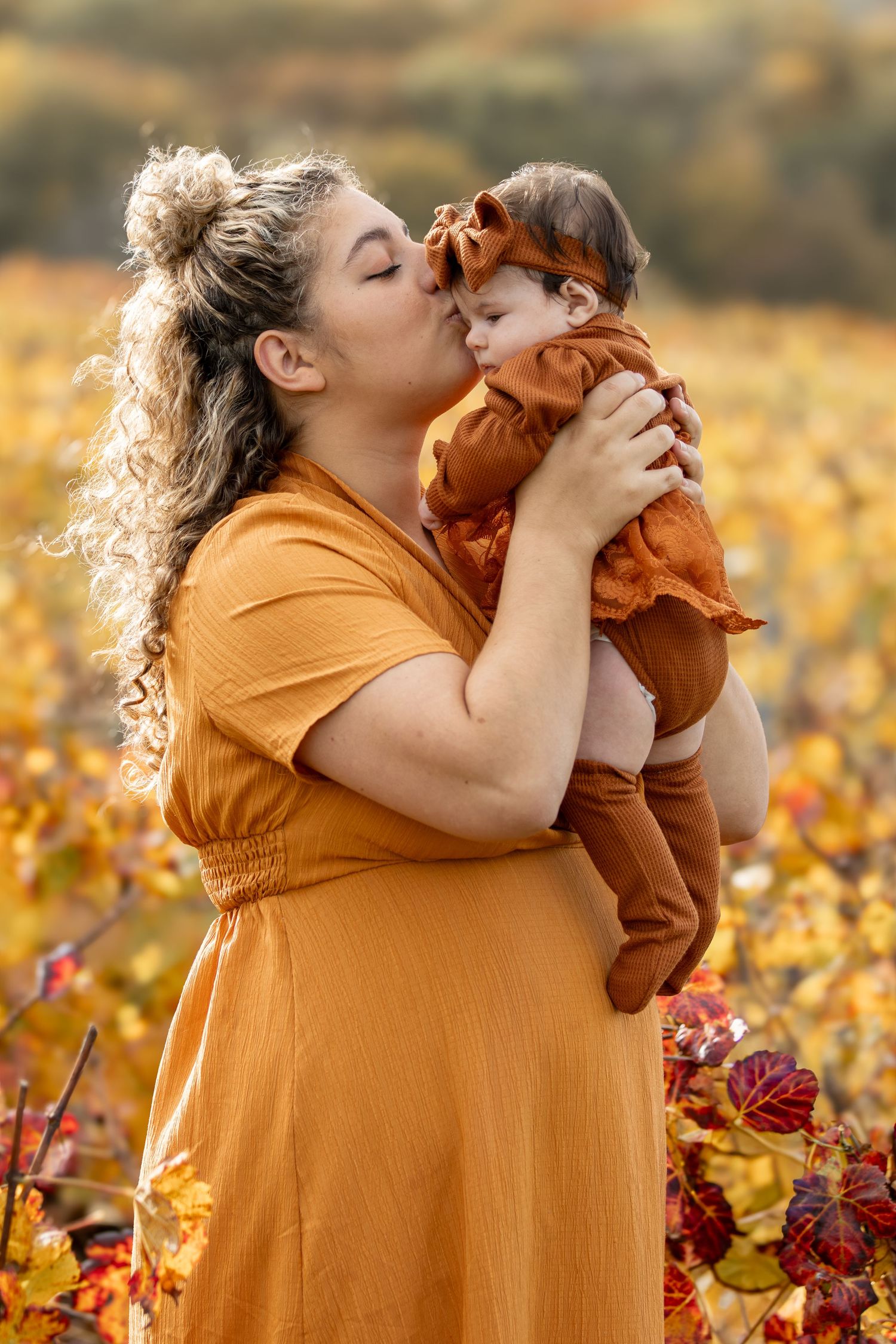 Mother in mustard dress kissing baby in rust romper in autumn vineyard.