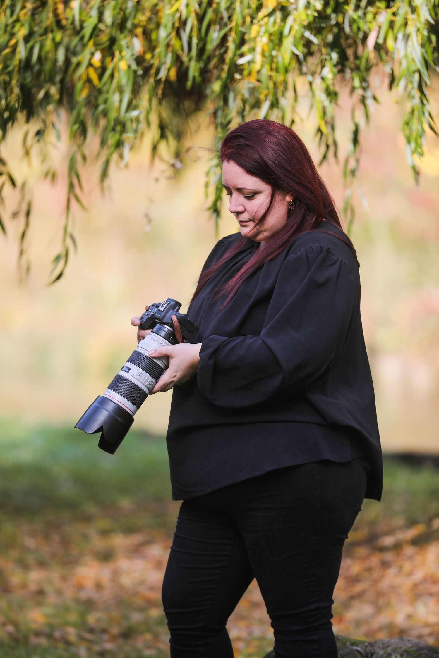 Female photographer reviewing settings on professional DSLR camera with telephoto lens under willow tree outdoors.
