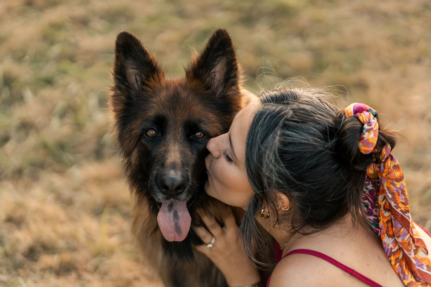 A young woman with a colorful hair scarf kisses her dark German Shepherd dog outdoors in a field.