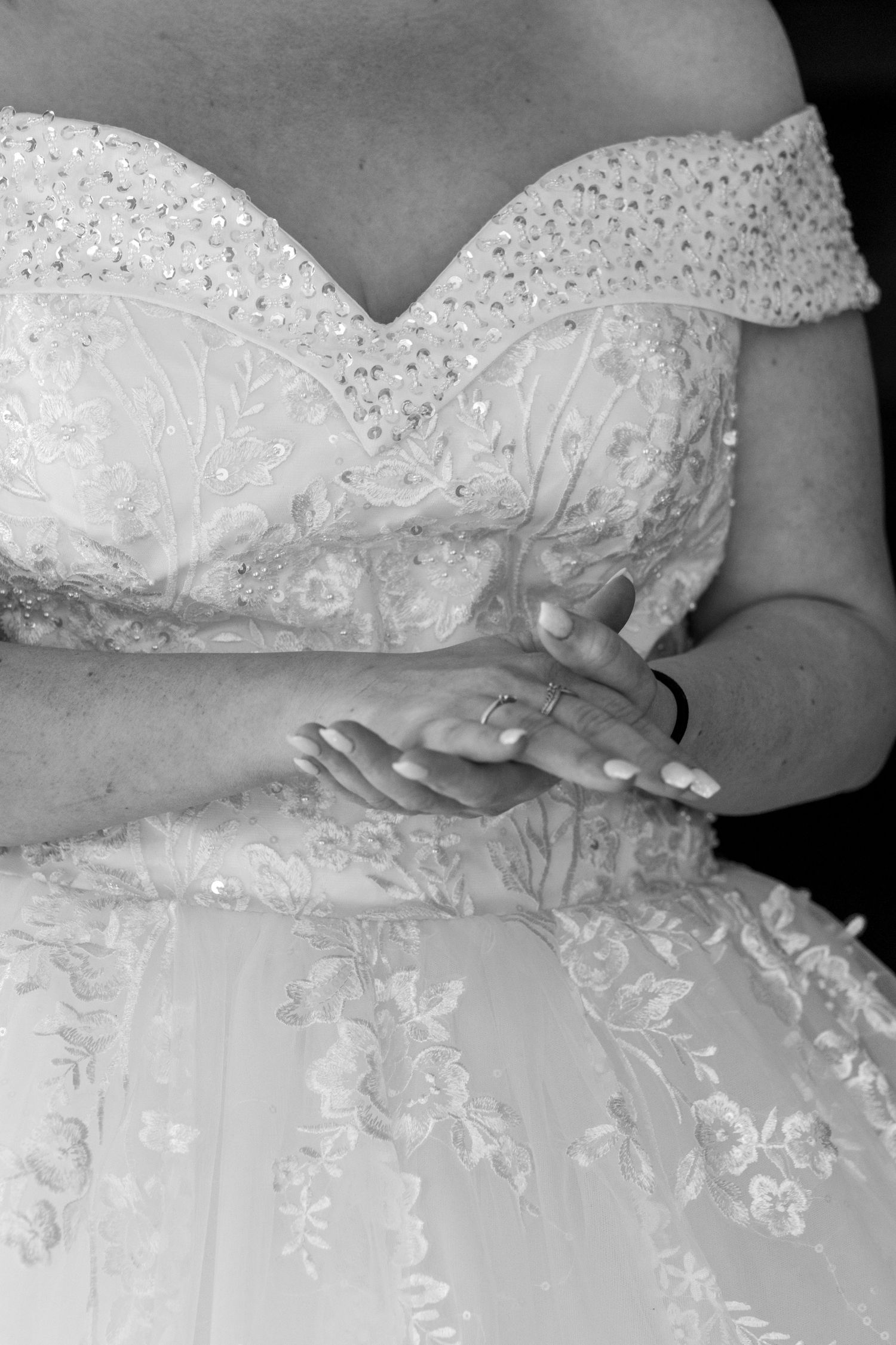 Black and white photo of a bride in a sequined off-shoulder wedding gown, hands clasped showing a wedding ring.