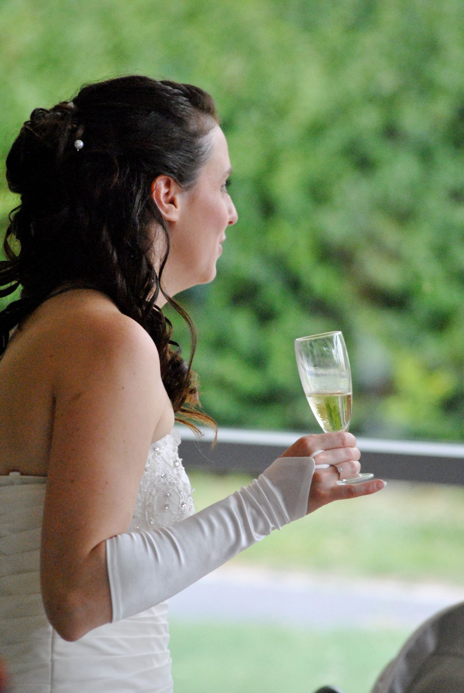 Beautiful bride in white strapless gown holds champagne glass outdoors.