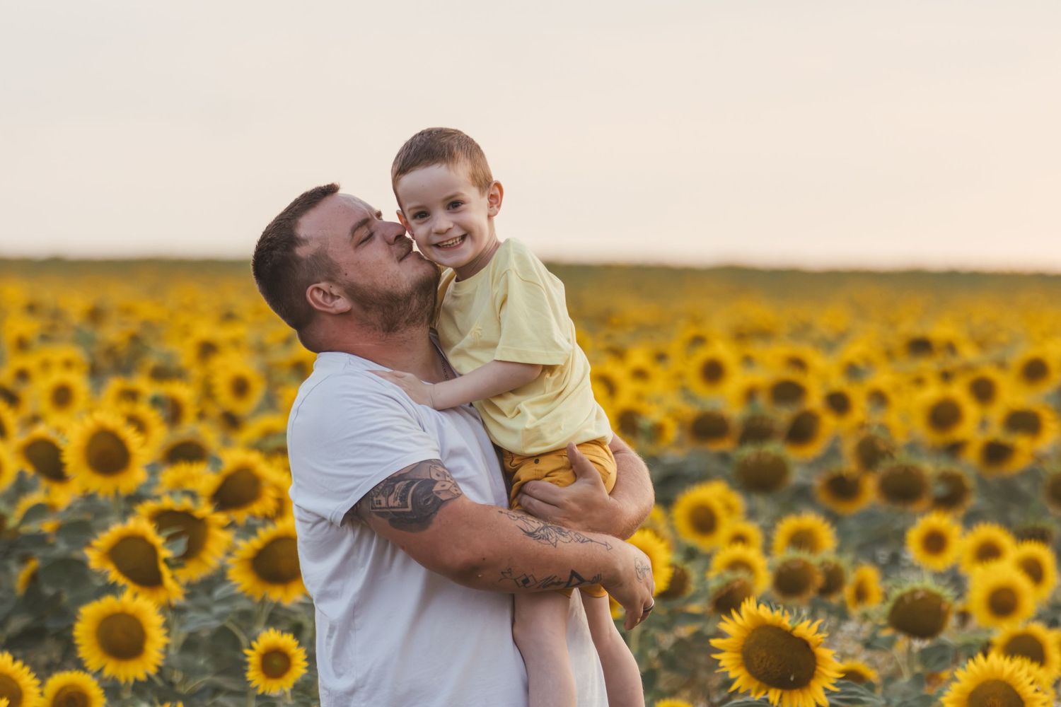 A father kisses his young son in a vibrant sunflower field at sunset.