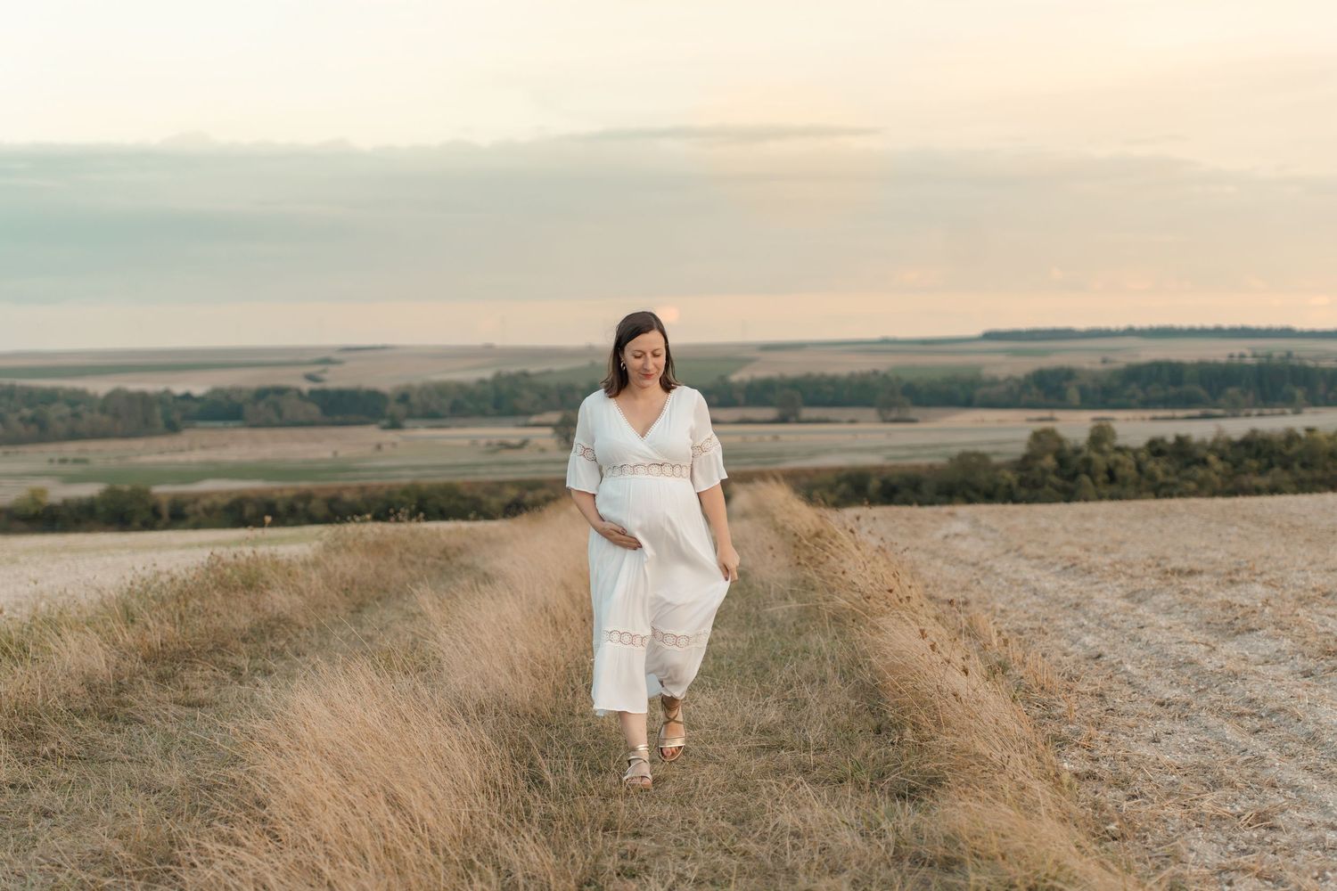 Pregnant woman in white dress walking through golden field at sunset with scenic countryside views.