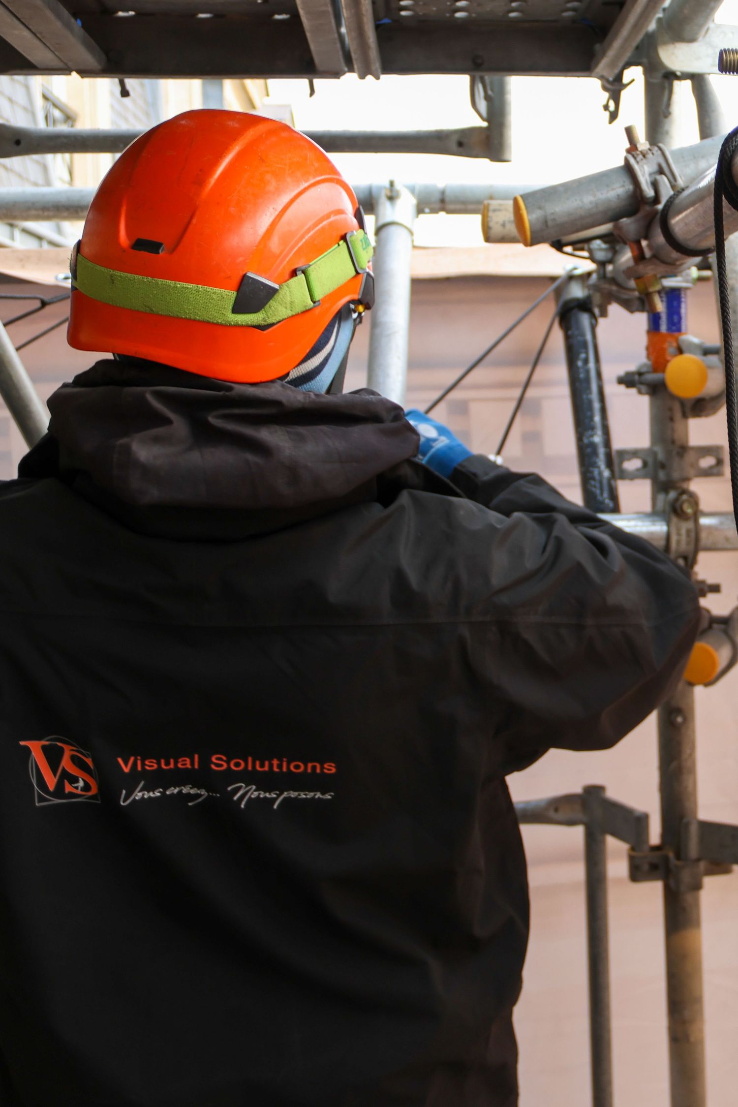 Worker in Visual Solutions jacket and orange safety helmet inspects scaffolding pipes at a construction site.