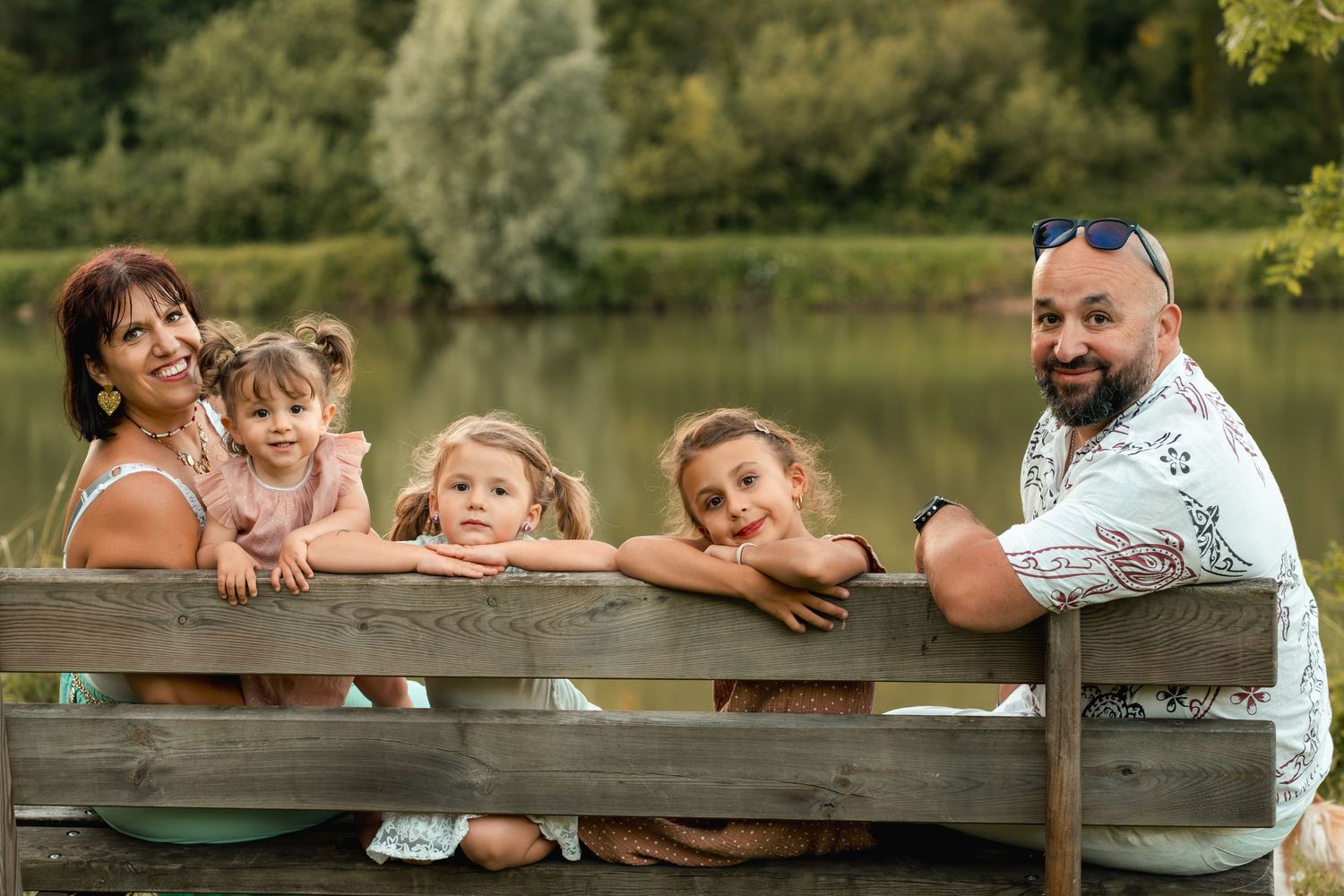 Happy family with three young girls posing on a wooden dock by a scenic lake on a sunny day.