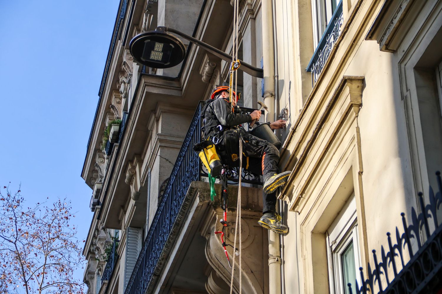 Worker in safety harness performing maintenance on ornate building facade with ropes and equipment in urban setting.