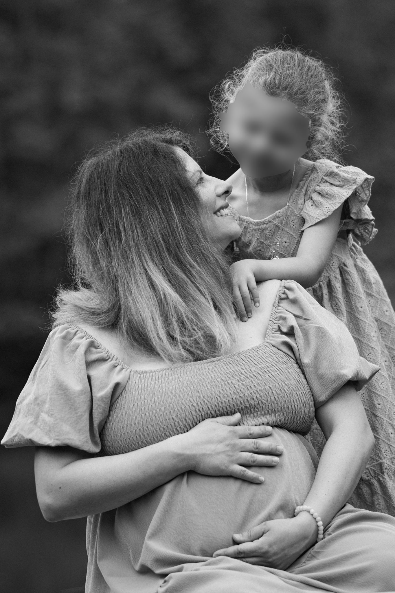 Black and white photo of a smiling pregnant woman holding a young girl outdoors.