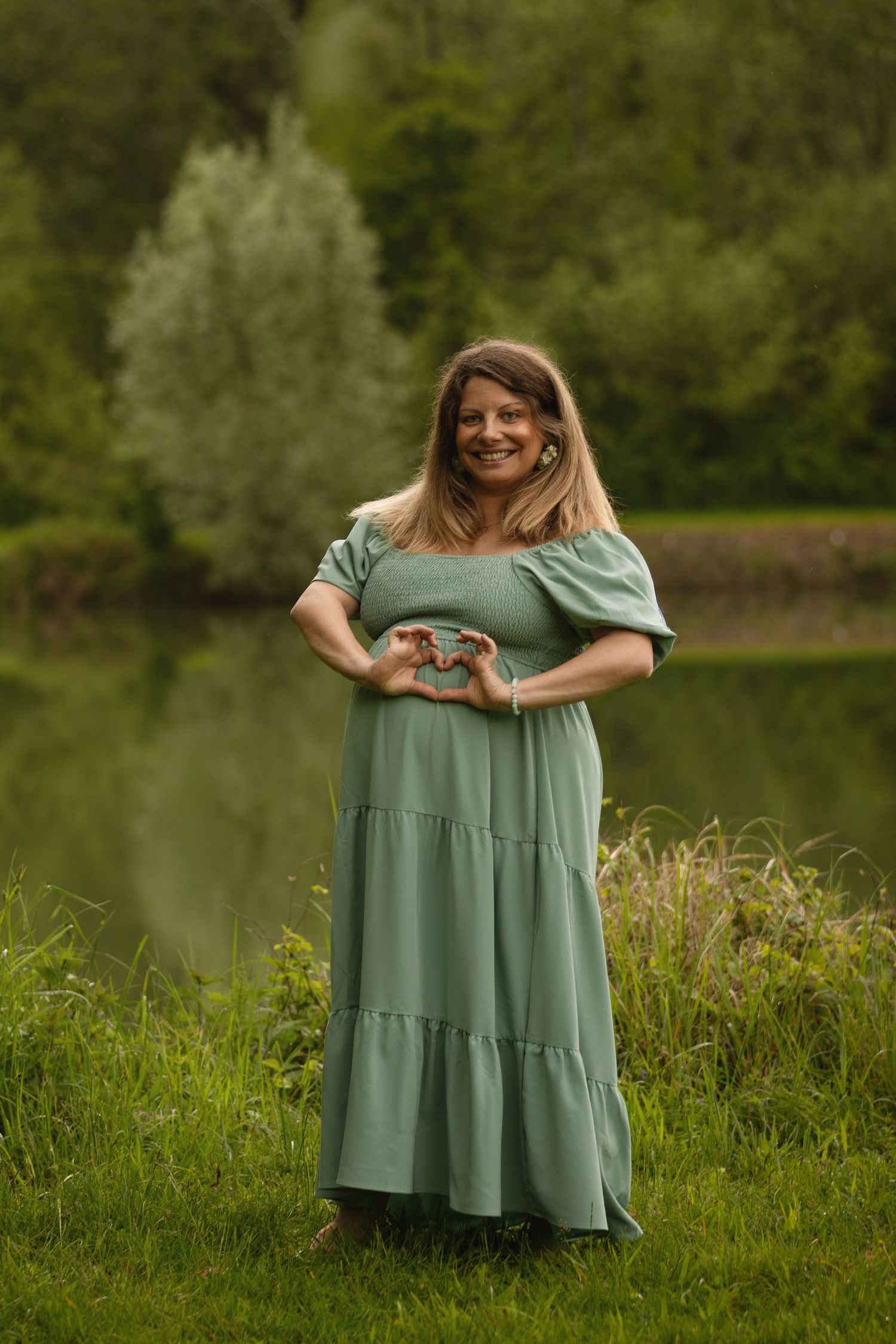 Smiling woman in sage green off-shoulder dress making heart shape with hands, standing by a serene pond.