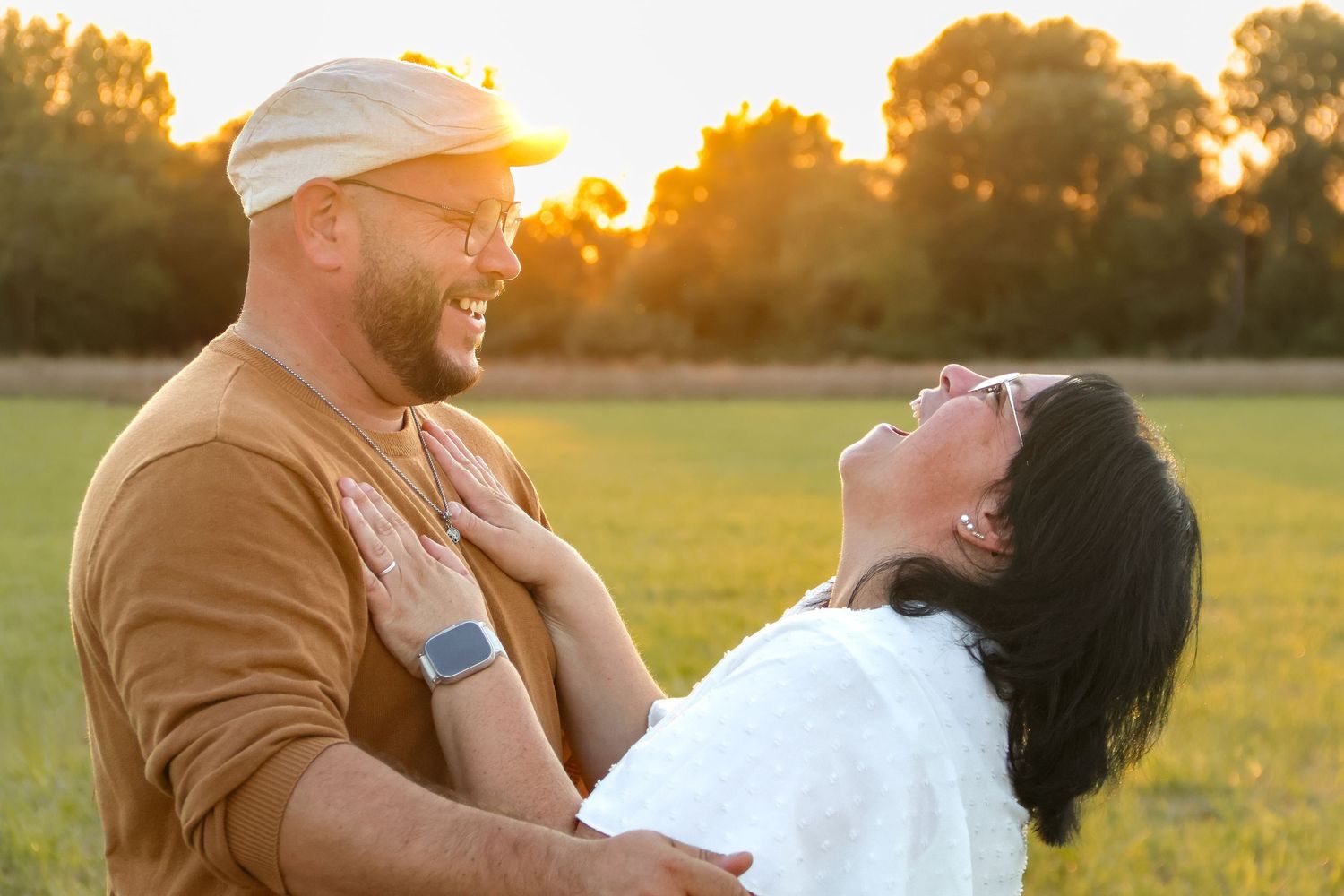 Happy couple laughing together in a sunny green field at golden hour, sharing a joyful romantic moment outdoors.