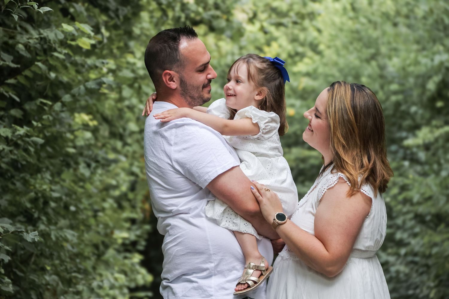 A couple holding their smiling toddler daughter outdoors surrounded by lush green foliage.