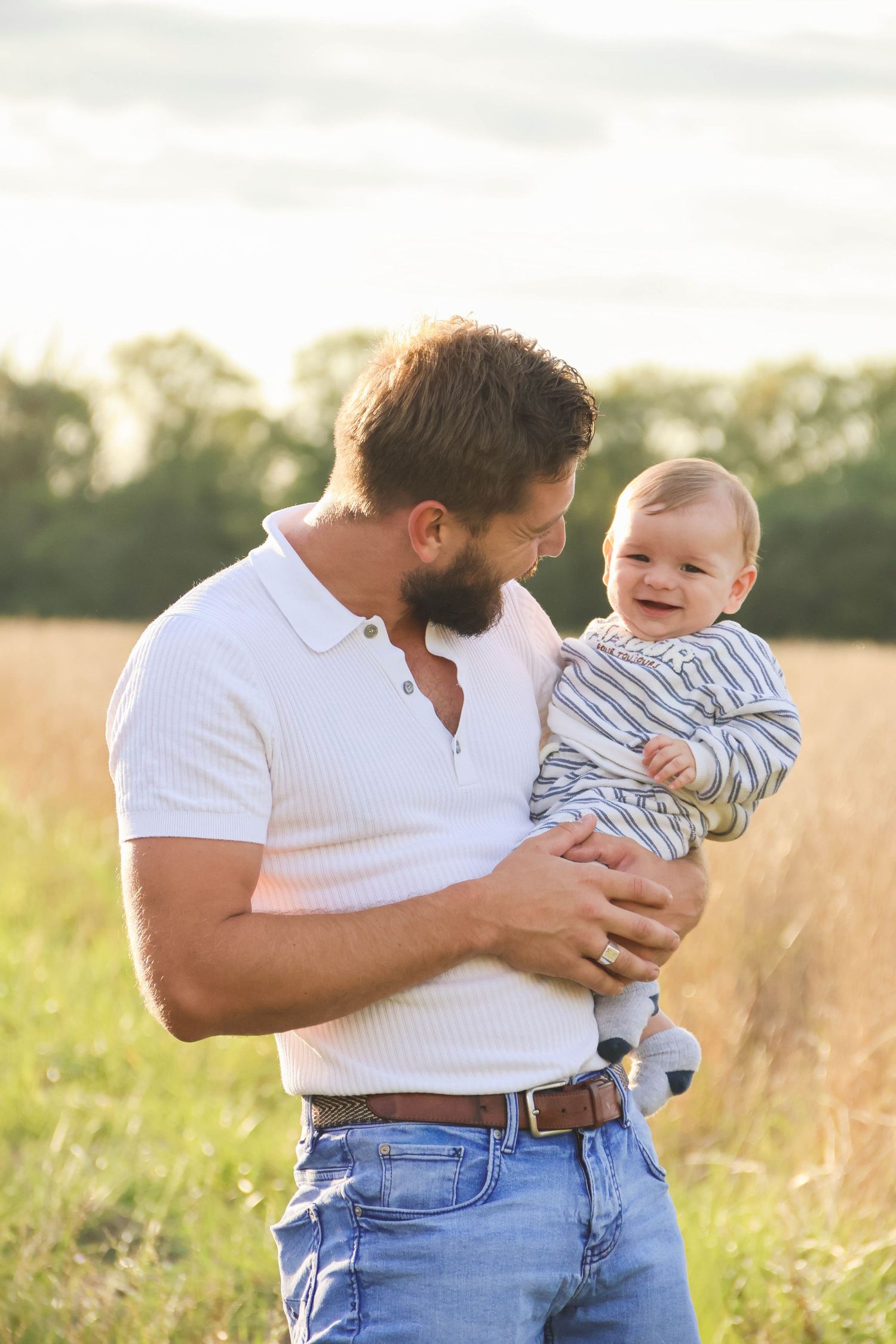 Séance famille — papa barbu en polo blanc tenant son bébé souriant dans un champ doré, photographe famille Aisne Guignicourt.