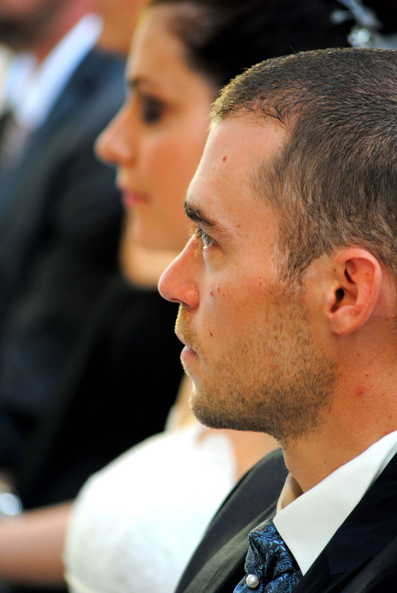 A groom in profile with short hair and blue tie attentively listening during a wedding ceremony.