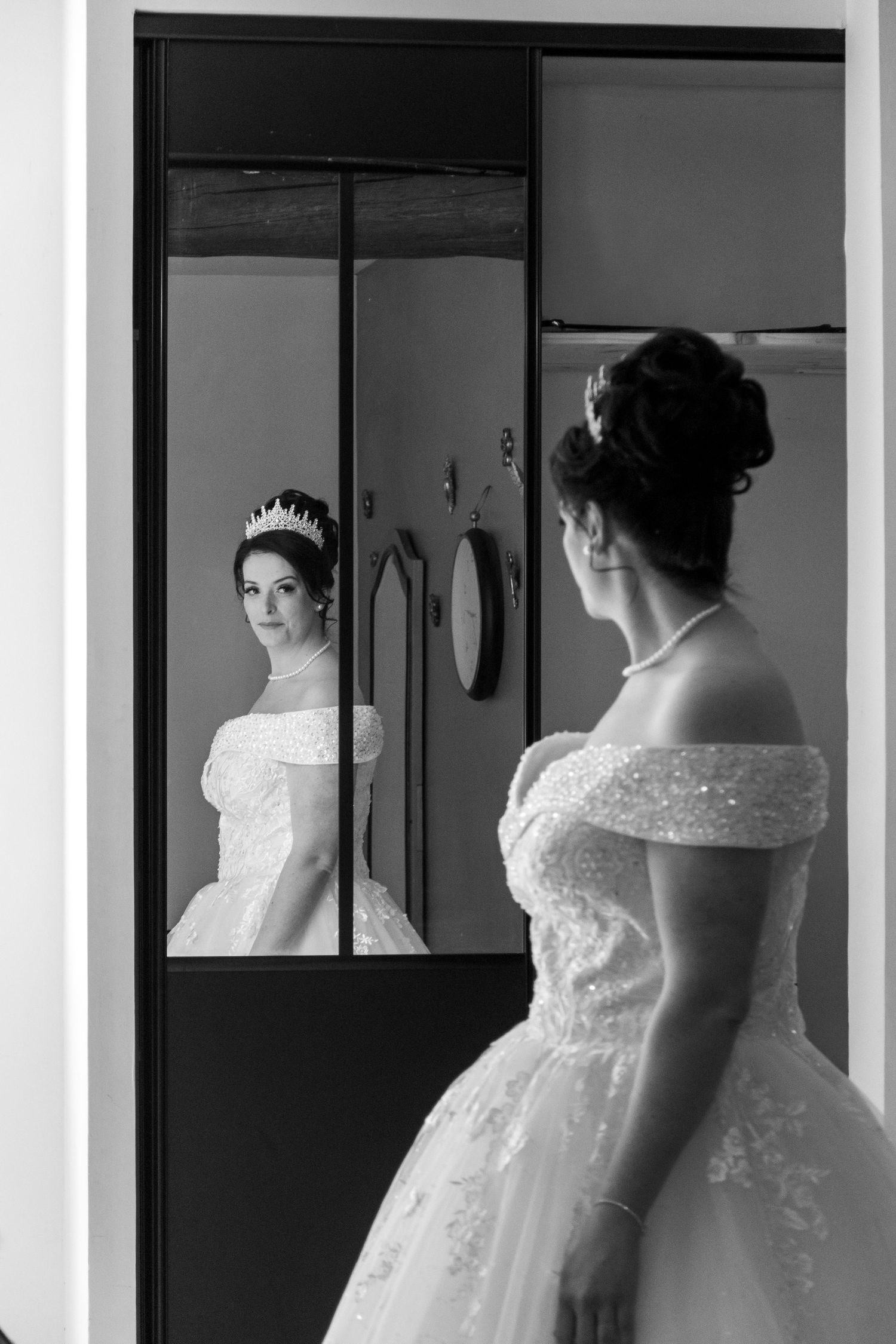 Black and white photo of a bride in a white gown and tiara admiring her reflection in a mirror.