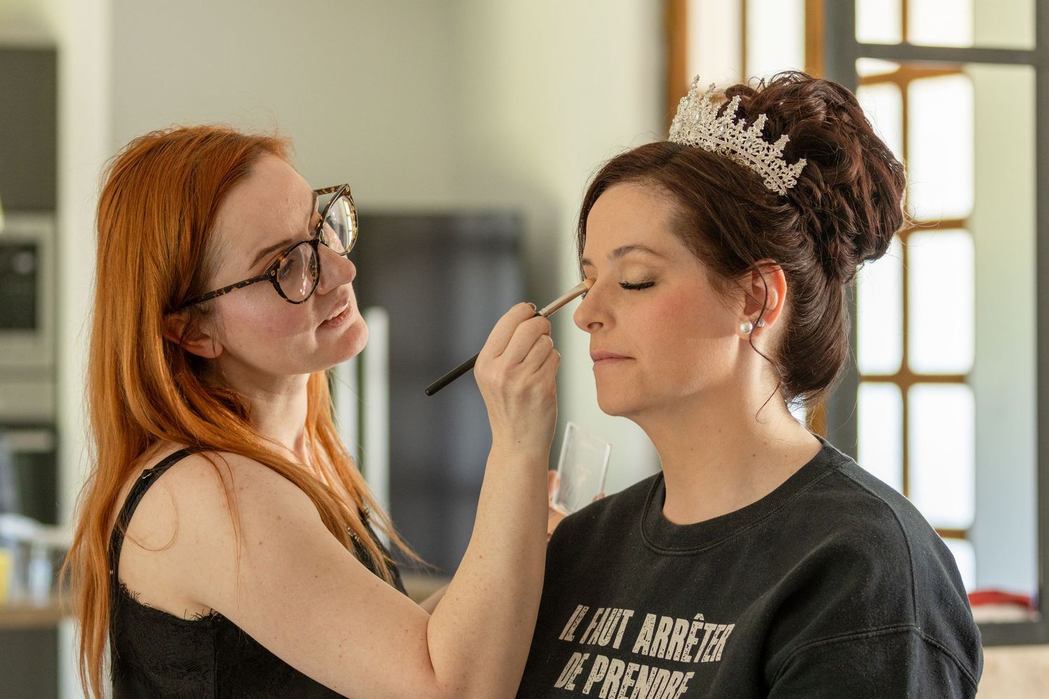 A makeup artist applying eye makeup to a bride wearing a tiara and black t-shirt in a bright room.