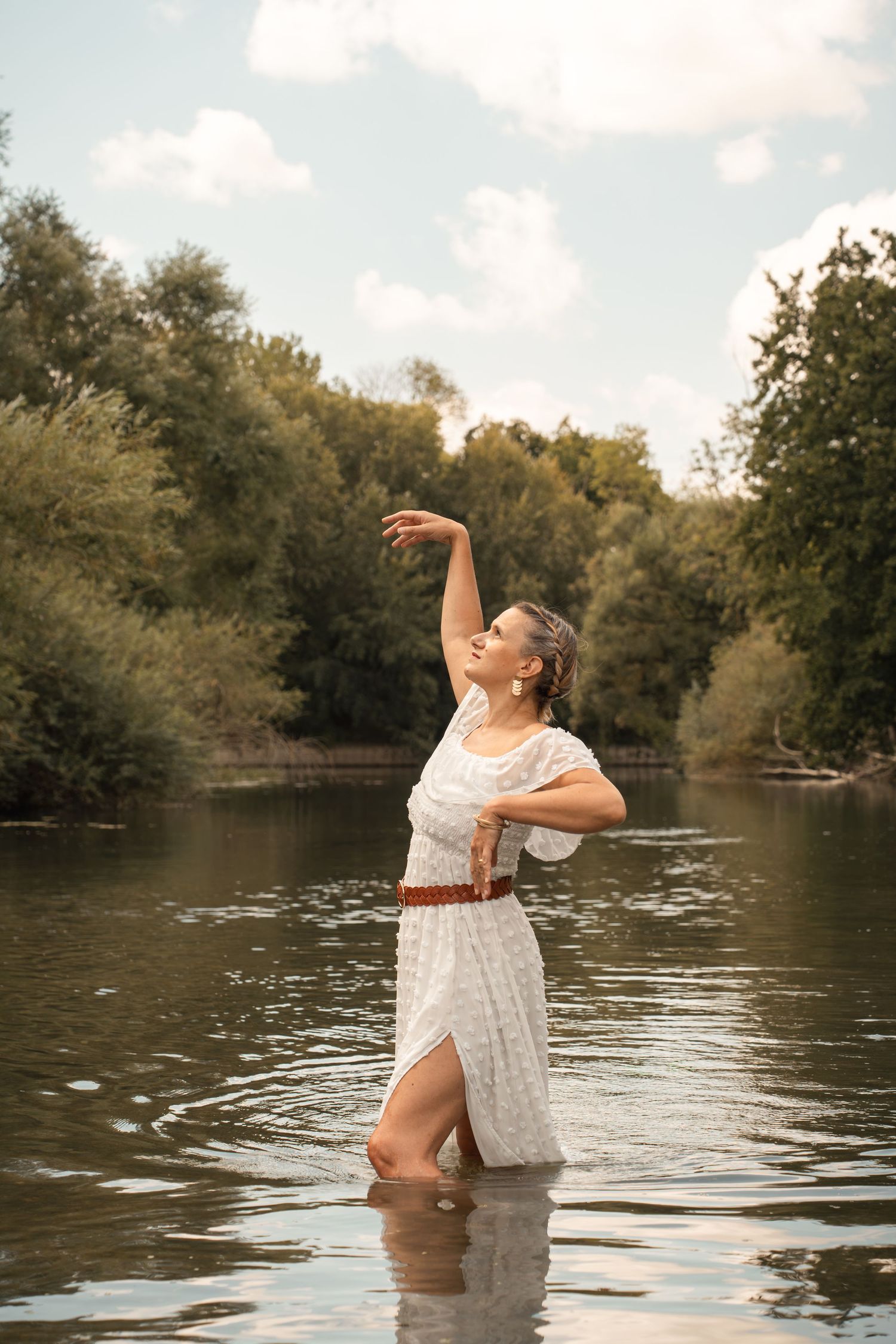 Woman in white dress with brown belt standing in river water, arms raised gracefully, surrounded by lush green trees.
