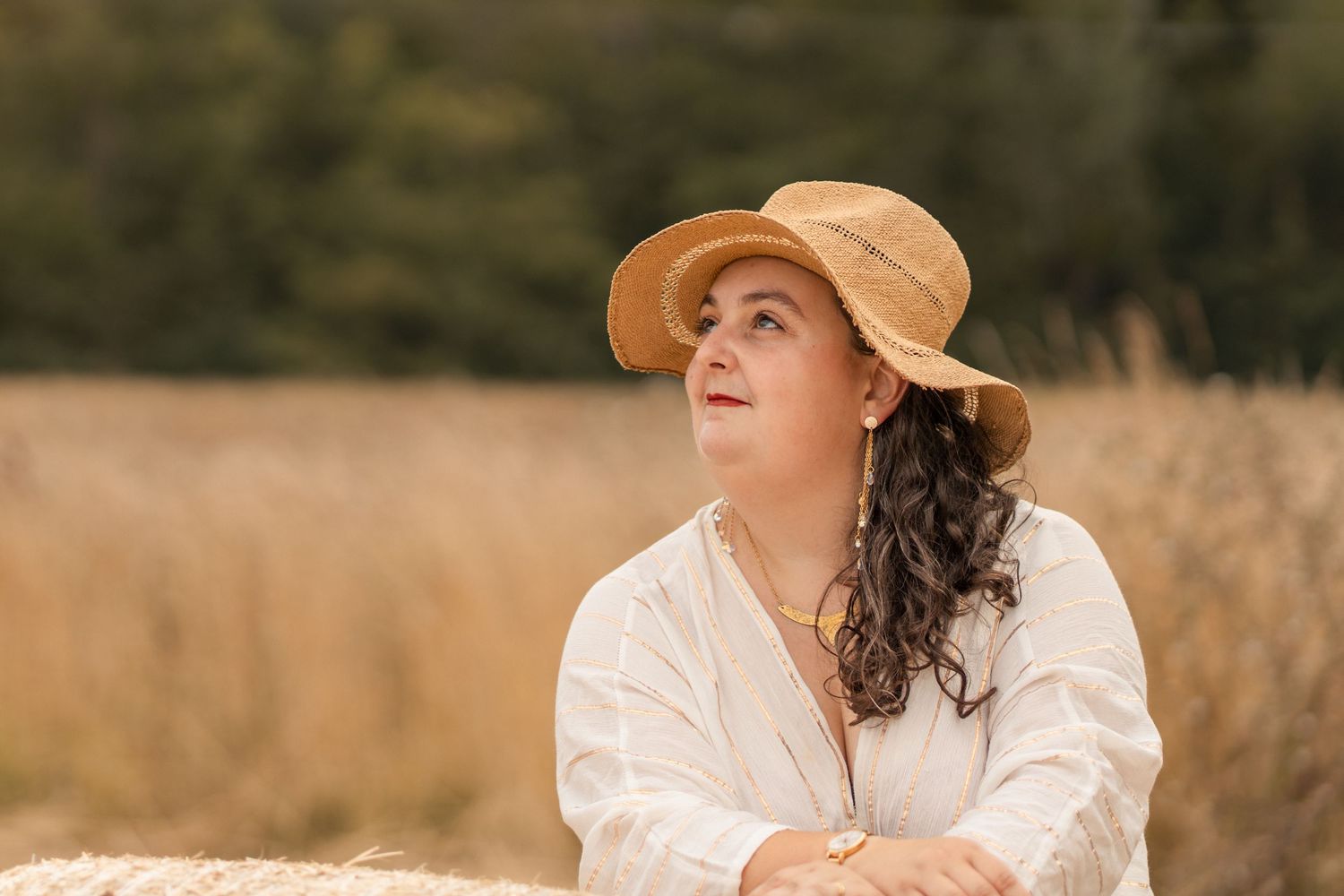 Woman in straw hat and white blouse leaning on hay bale in golden wheat field on sunny day.