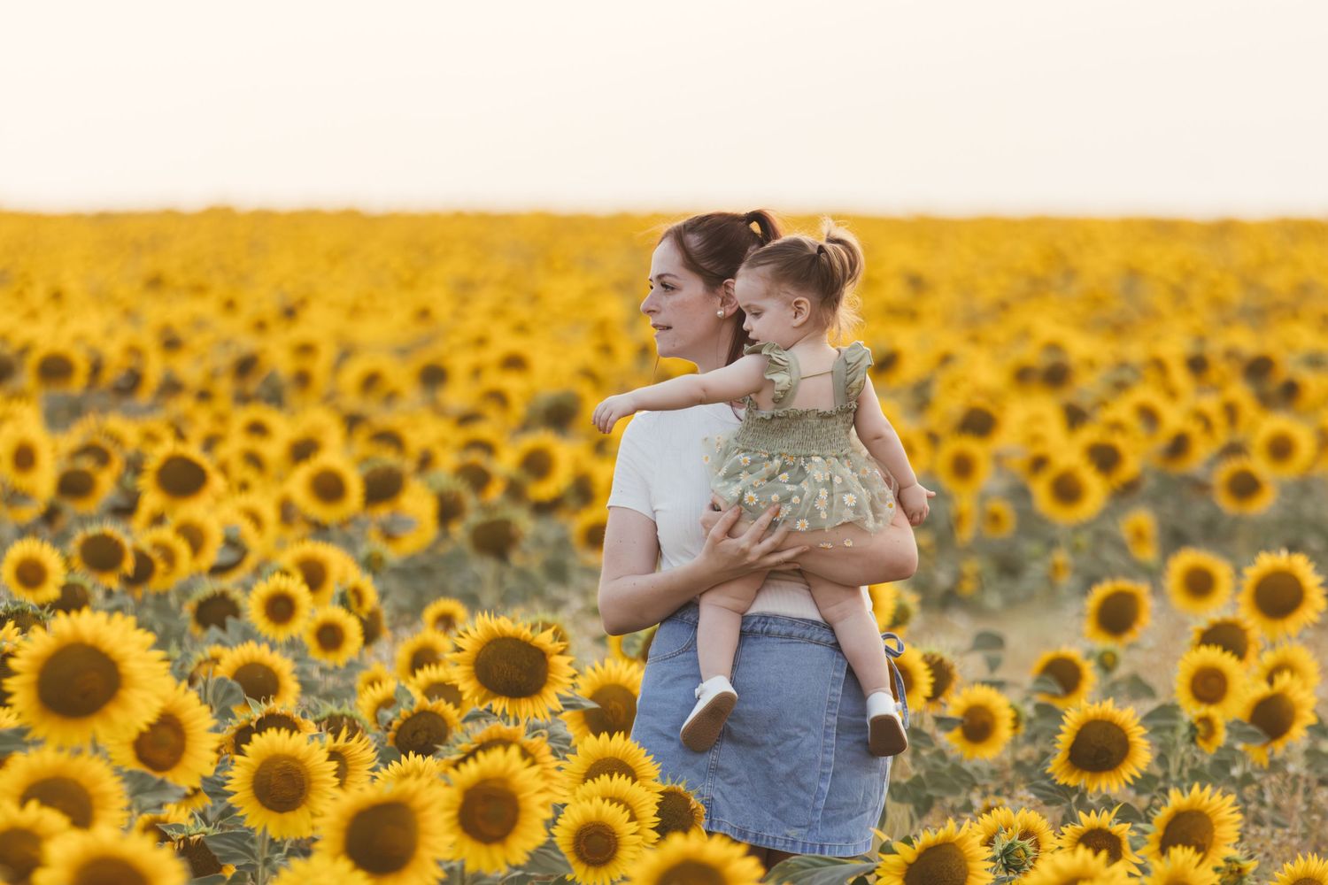 Mother holding baby girl in sunflower field at sunset, surrounded by bright yellow blooms.