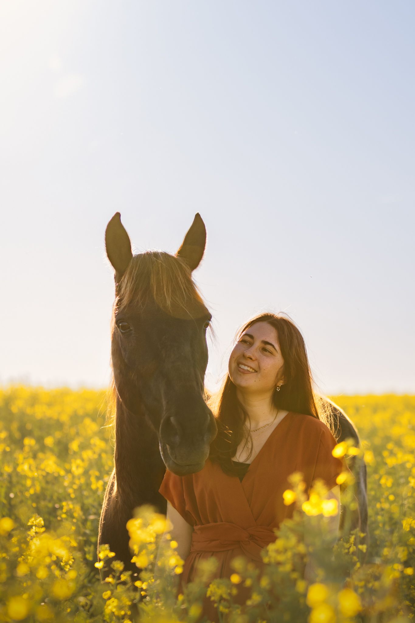 Jeune femme souriante en robe rouge debout à côté d'un cheval brun foncé dans un champ de fleurs sauvages jaunes ensoleillé.