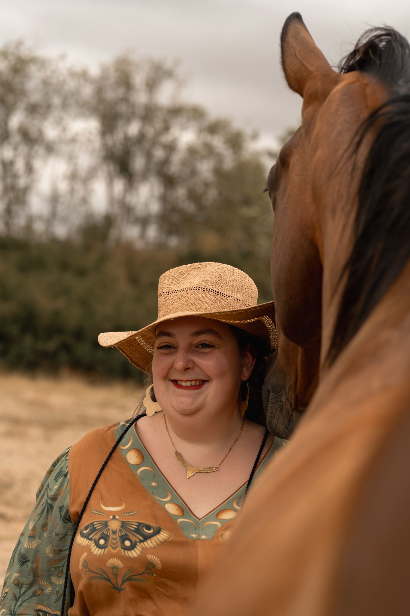 Smiling woman in cowboy hat and floral top standing beside a chestnut horse outdoors.
