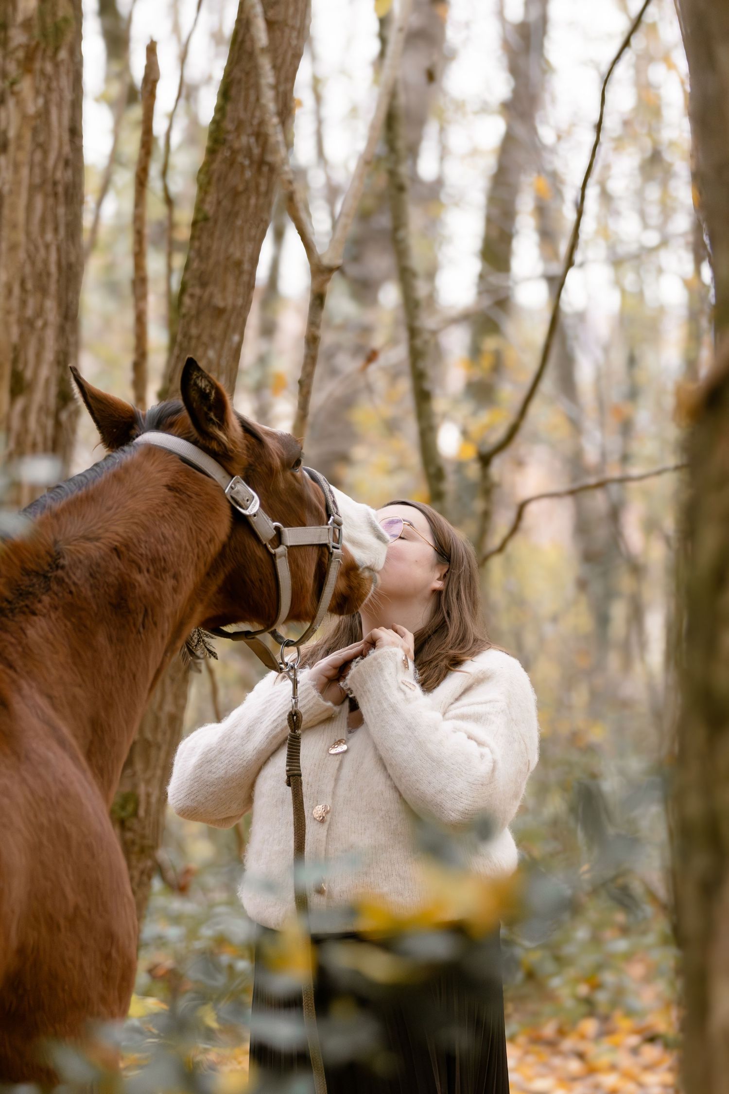 Young woman affectionately kissing a brown horse with white blaze in fall woodland.
